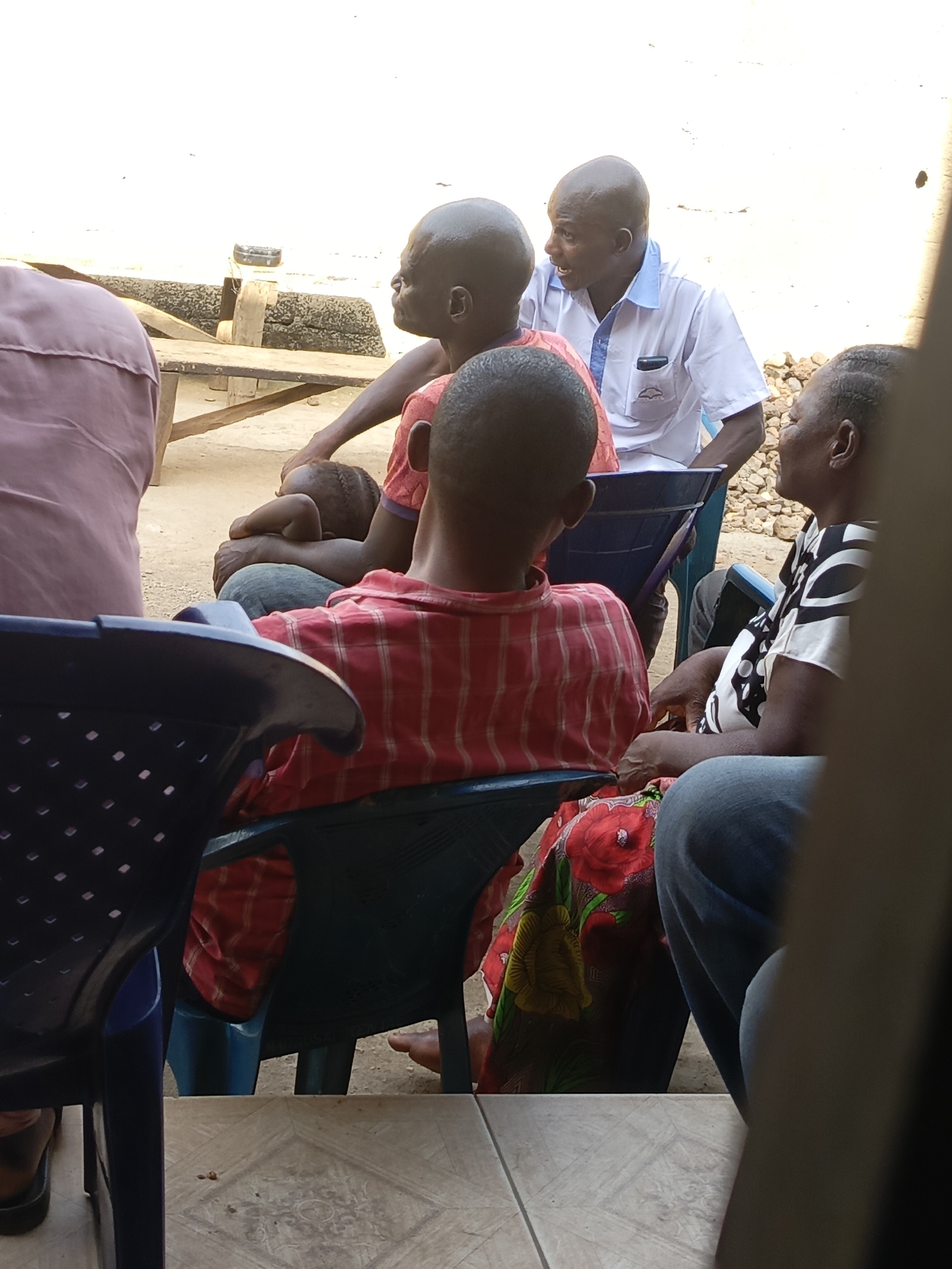 Several people are gathered outside in Akwanga, Nigeria. One man in a white shirt and blue trim appears to be speaking to another man with a bald head. A child with braided hair rests on the lap of one of the men. A woman wearing a patterned shirt is present, as well as other individuals whose details are partially obscured by chairs. The gathering is taking place outdoors, with a table and some objects visible in the background. The setting is on a tiled surface.