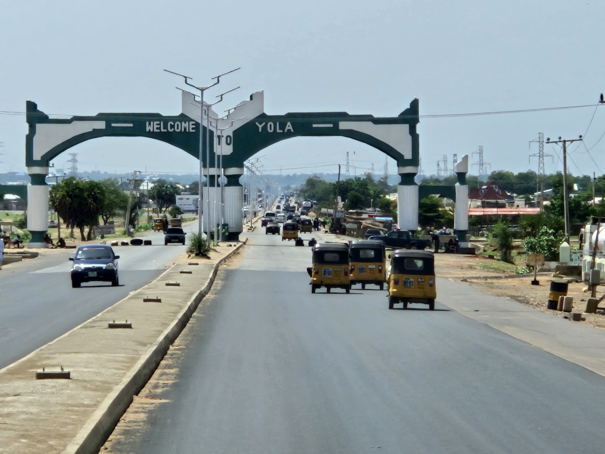 The image shows a wide asphalt road leading into the city of Yola, Nigeria, identified by a prominent green and white "WELCOME TO YOLA" archway spanning the road. A concrete median divides the road. Several yellow auto rickshaws (tuk-tuks) are traveling towards the viewer. Further in the distance, other vehicles, including cars and trucks, are visible on the road. On the right side of the road, there are objects such as barrels, blocks, and posts. Along the sides of the road are trees, bushes, buildings, and power lines. In the background, electrical towers are visible.