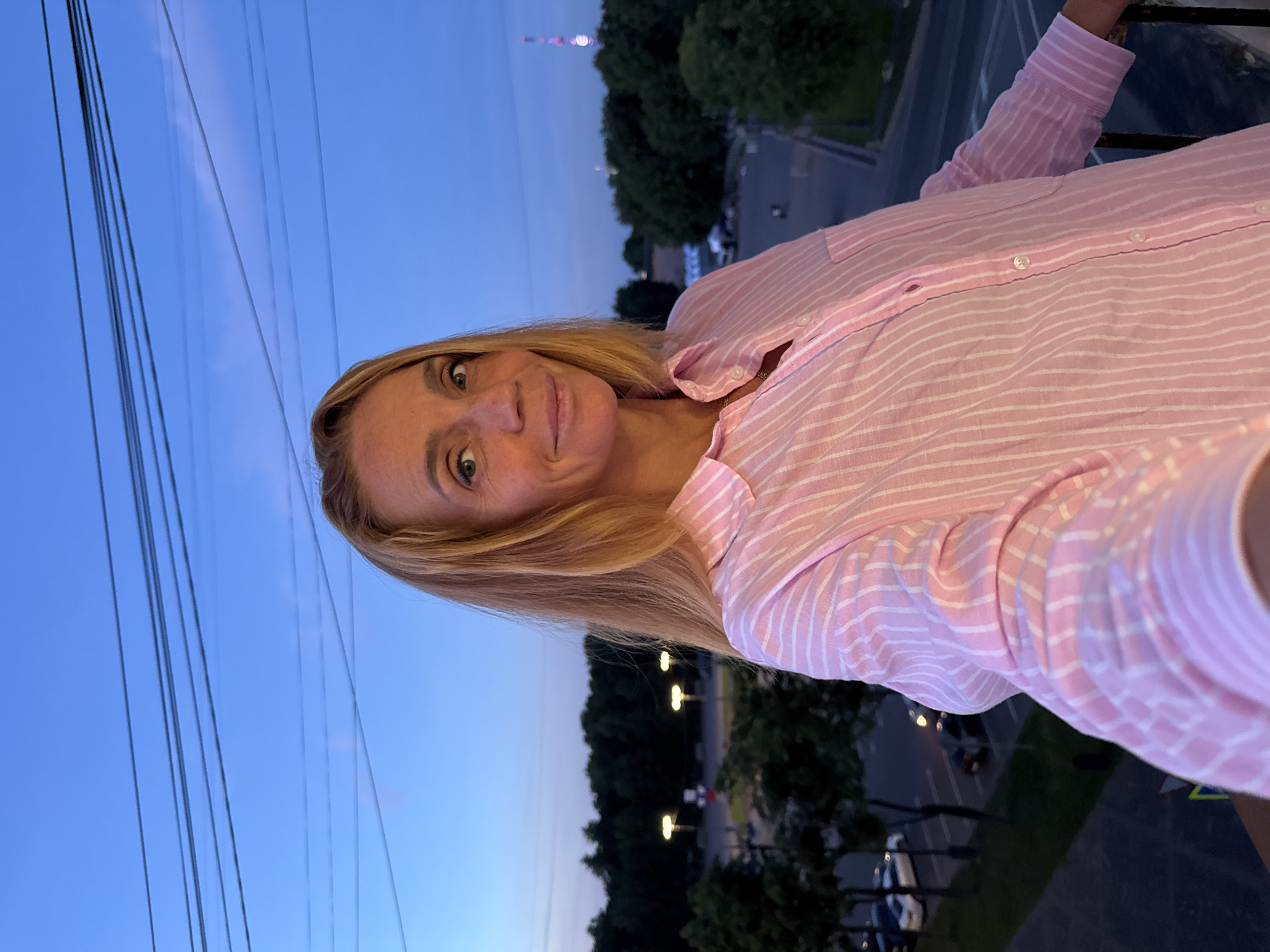 A young adult woman with blonde hair and blue eyes is pictured from the chest up, taking a selfie with a slight smile. She is wearing a pink and white striped button-down shirt and is standing outdoors on a balcony. Behind her, the sky is a deep blue, indicating it is dusk. There are power lines visible across the top of the frame. In the background, a cityscape is visible with trees lining a street, streetlights, and buildings. A tall tower with a light at the top can be seen in the distance. The overall impression is that the photograph was taken in Veliky Novgorod, Russia, during the evening.