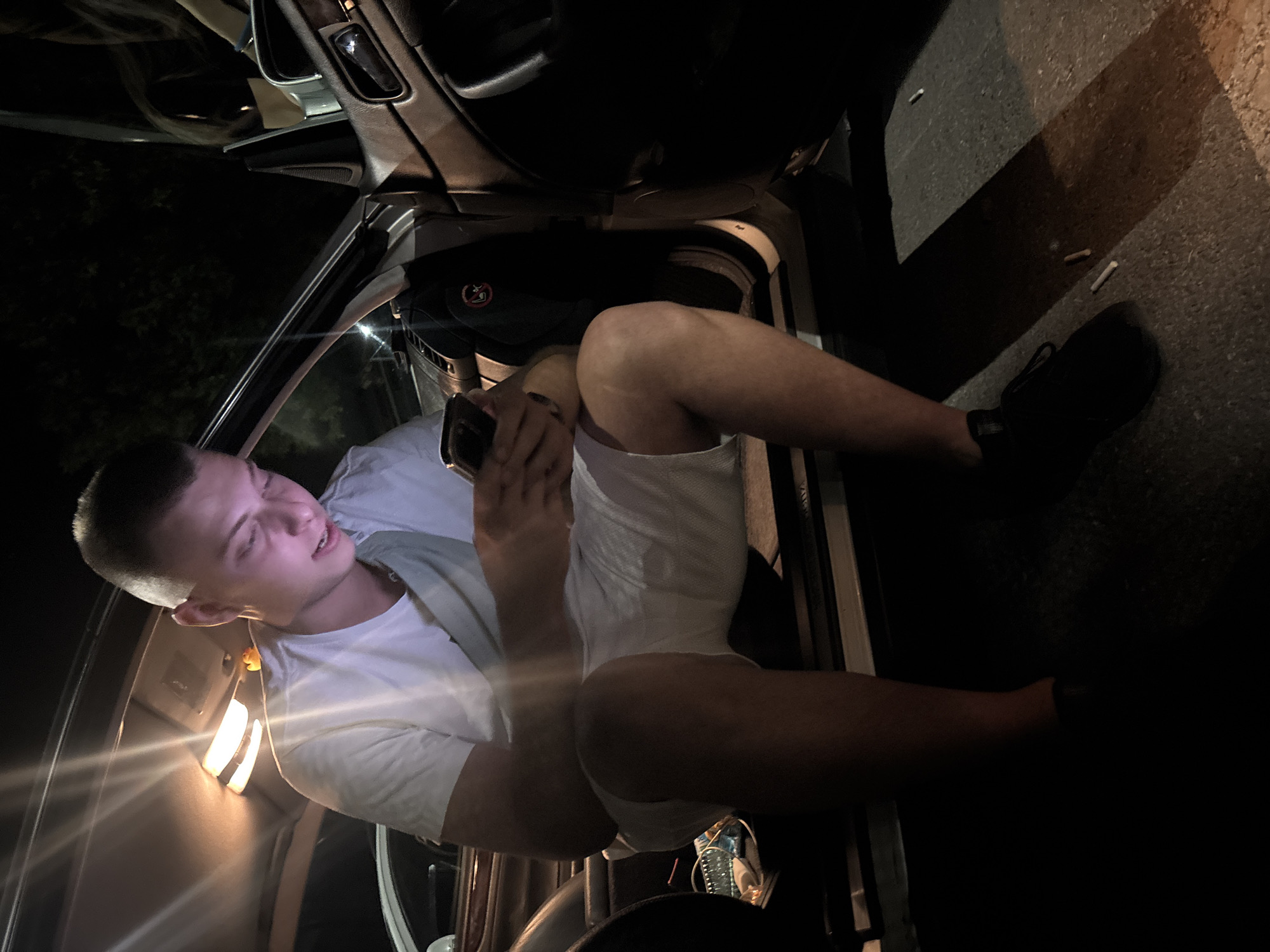 A young man, appearing to be in his early twenties, is seated in the open driver's side of a car at night. He is wearing a white t-shirt and white shorts, and black sneakers. He is looking down at his smartphone, which he is holding in both hands. His left leg is bent, with his knee pointing upwards, and his right leg is also bent, with his foot resting on the ground outside the car. The interior of the car is lit by an overhead light, and the exterior is dark. Two cigarette butts are visible on the ground near the car door. The image appears to have been taken on a street or parking lot. The man's clothing and the presence of the car suggest he is outdoors. The darkness and overhead lighting are consistent with a nighttime scene in an urban or semi-urban environment.