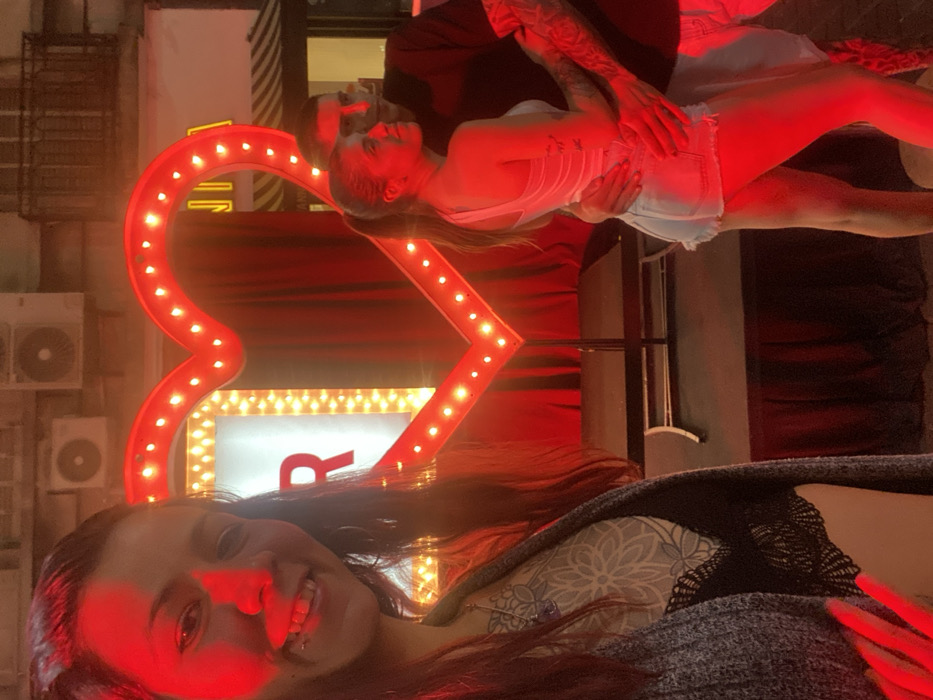 The image captures a vibrant night scene illuminated by red light, featuring two women and a man. The original image appears to be rotated 90 degrees counter-clockwise; when viewed correctly in landscape orientation, the scene unfolds as follows:
In the left-center of the frame, a man and a woman are engaged in a close embrace or dance pose, bathed in intense red light. The man stands behind the woman, holding her around the waist. His arms are visibly tattooed with intricate designs. The woman is wearing a light-colored, possibly white, strapless top and light-wash denim shorts. Her head is tilted back, and she appears to be looking towards the man, or slightly upwards. Her leg is extended, adding to the dynamic pose.
Behind this couple, a large, prominent heart-shaped sign with numerous red light bulbs creates a romantic backdrop. Below the heart, a rectangular marquee sign with yellow lights is visible, featuring a large red letter "R" prominently displayed.
To the far left of the image, in the background, parts of a building or wall are visible, with several outdoor air conditioning units mounted on it. The immediate background behind the couple and the signs is dark, suggesting a stage curtain or a dark wall, further emphasizing the illuminated figures.
In the right foreground of the image, a different woman is captured from the chest up, smiling broadly towards the camera. Her face is strongly lit by the red light, highlighting her features and bright smile. She has long, dark hair and is wearing a dark grey, possibly knitted, top over a lighter, patterned garment.
The overall mood is lively and romantic, primarily due to the dominant red lighting and the suggestive poses of the central couple. The woman in the foreground conveys a sense of happiness and direct engagement with the viewer. The scene appears to be an outdoor evening setting, perhaps at an event or a venue with decorative lighting.