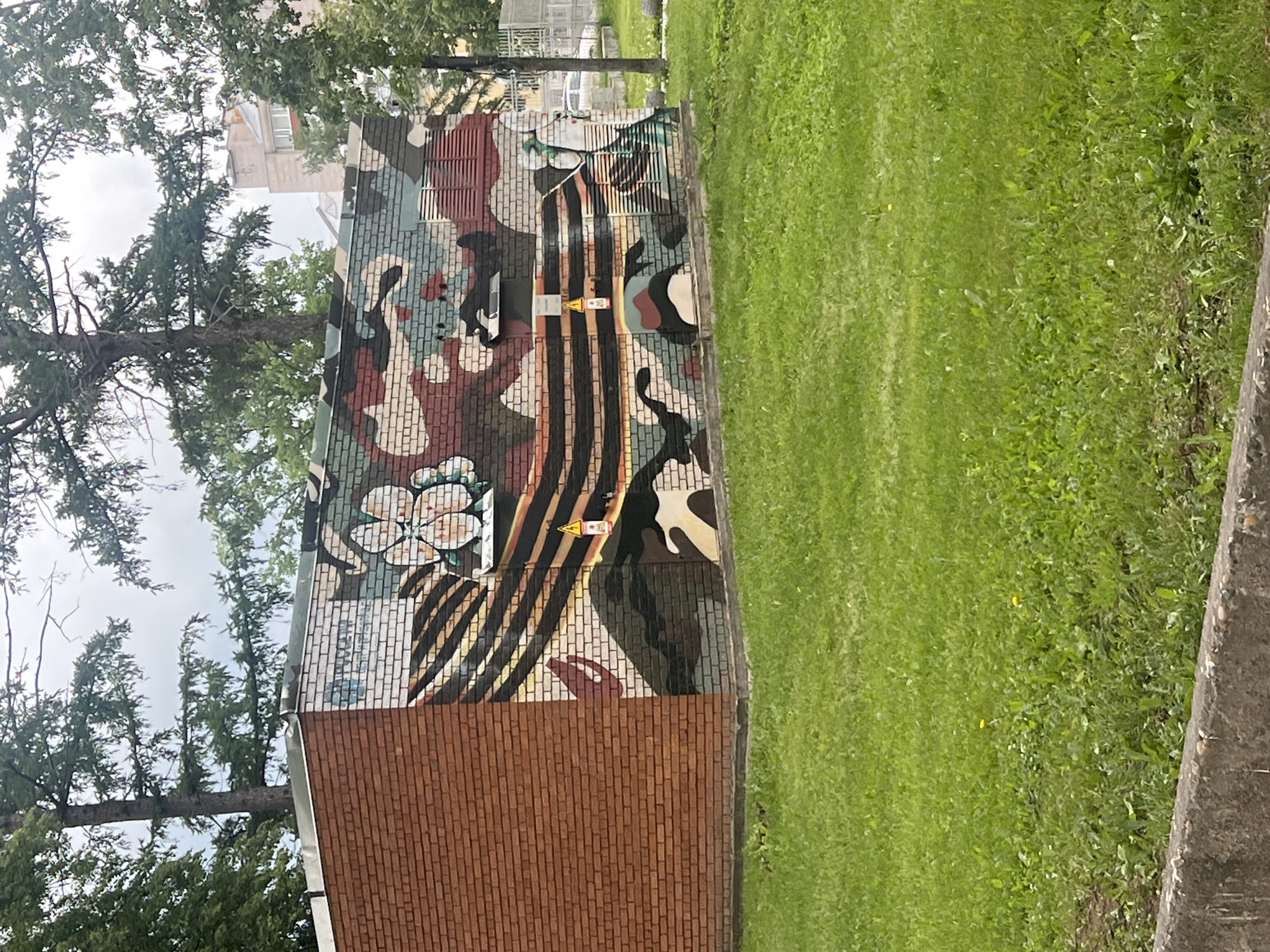 The image captures an outdoor scene in Votkinsk, Russia, during what appears to be a bright or overcast daytime. The main subject is a low-rise brick structure, possibly a utility box or a small building, which is partially covered by a vibrant mural. The lower portion of this structure is made of plain red bricks.
The mural itself is painted on the upper part of the structure and features a complex design. It incorporates a military-style camouflage pattern with various shades of green, brown, black, grey, and white/cream. A prominent white, multi-petaled flower, resembling a poppy or similar blossom, is depicted on the left side of the mural. Horizontally across the center, a distinctive ribbon with alternating orange/gold and black stripes, characteristic of the Russian St. George ribbon, is painted. Several yellow triangular warning signs with black pictograms, likely indicating electrical hazards, are affixed to the mural.
To the left of the structure and extending into the background, tall evergreen trees, possibly pines or firs, stand against a light sky. A light-colored building is partially visible through the trees in the distance. The ground adjacent to the structure on the right side of the image is covered in lush, well-maintained green grass. A grey concrete curb or border runs along the edge of the grassy area on the far right. No people or specific activities are visible in the image.