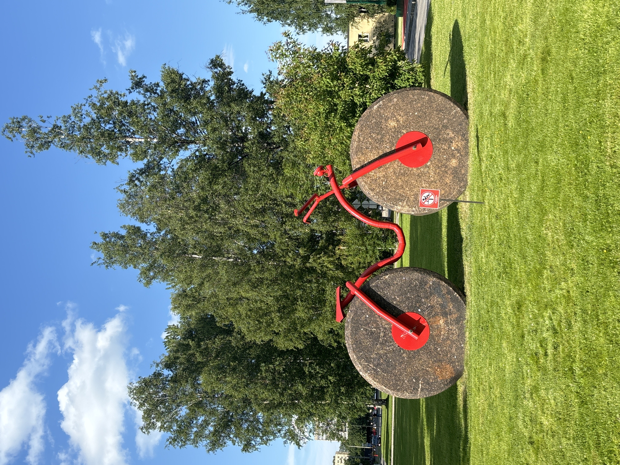 The image shows a large, red metal sculpture of a bicycle with two massive millstones as wheels. The sculpture is situated in a well-maintained grassy park with trees in the background. A clear blue sky with scattered white clouds forms the backdrop. A small red and white sign with a pictogram of a person falling is planted in the grass near the front wheel of the sculpture. In the distance, there are buildings and a road with some vehicles. The sculpture is located in Saint Petersburg, Russia, at the entrance to the Alexander Park, which is part of the Peter and Paul Fortress complex. The time of day appears to be daytime, with bright sunlight casting shadows on the grass.