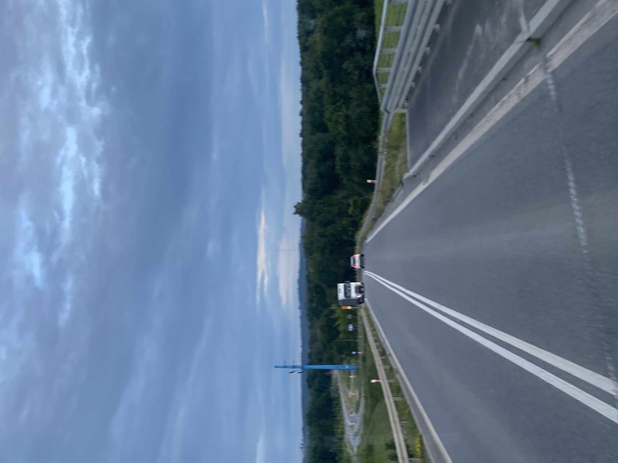 The image shows a road scene from the perspective of a vehicle driving on a highway. The road is paved with asphalt and has double white lines marking the lanes. There are guardrails on the sides of the road. In the distance, a white truck and a white car are visible, both driving in the same direction as the camera. The landscape on either side of the road consists of green trees and vegetation. The sky is overcast with dark blue clouds. A distinctive blue utility pole with multiple arms is visible to the left of the road, along with a curved exit ramp. The location is identified as Podłęże, Poland.