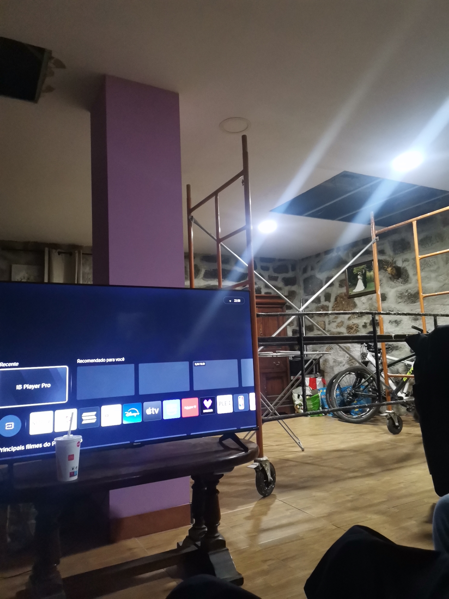 Here is a description of the image:
**Setting:** The photo appears to be taken indoors, possibly inside a home. The room seems to be under renovation or construction, as evidenced by scaffolding. The walls are made of stone.
**Subjects:**
* The foreground is dominated by a large flat-screen TV displaying an interface with an "IB Player Pro" selection. There is also a paper cup with a straw on the table that the TV is sitting on.
* There's a bicycle in the middle-ground and someone is sitting down in the lower right corner.
**Objects:**
* Flat-screen TV.
* TV stand.
* McDonald's cup.
* Scaffolding.
* Bicycle.
* Artwork/decorations on the wall.
**Action/Interactions:** No explicit actions are taking place. The scene seems static, with someone watching TV in the middle of renovation work.
**Environment:** The environment is chaotic due to the scaffolding and renovation in progress. The natural light appears to be coming in from an opening above. The room has stone walls, giving it a rustic appearance. The room appears well-lit.