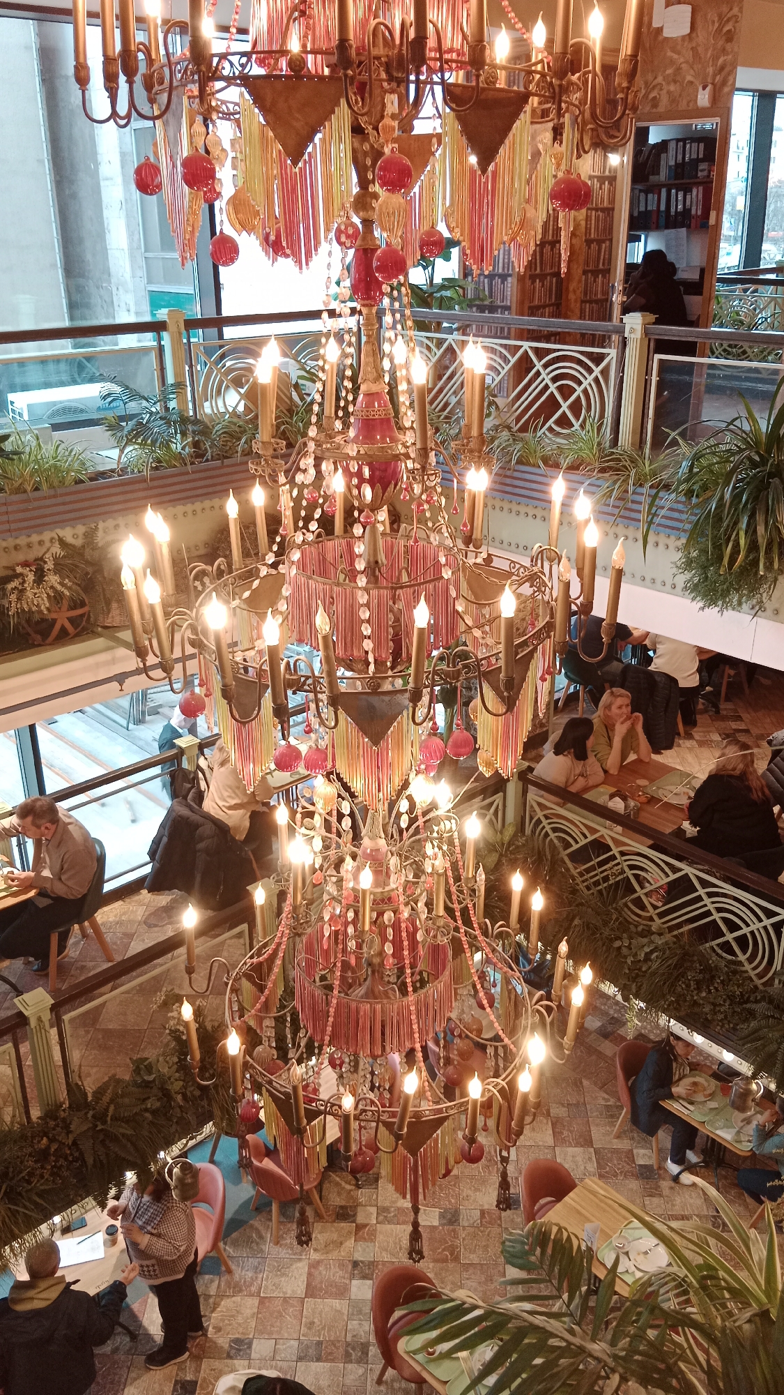 The image shows a multi-level restaurant interior, likely taken from an elevated perspective, focusing on a large, ornate chandelier. The chandelier has multiple tiers, each adorned with faux candles, beads, and colorful tassels. The restaurant features a tiled floor in a geometric pattern. Tables and chairs are arranged throughout the space, with patrons seated and engaged in conversation or dining. A glass railing separates different levels, decorated with plants and foliage. A bookshelf is visible in the background on an upper level. The location is Unknown, Unknown.