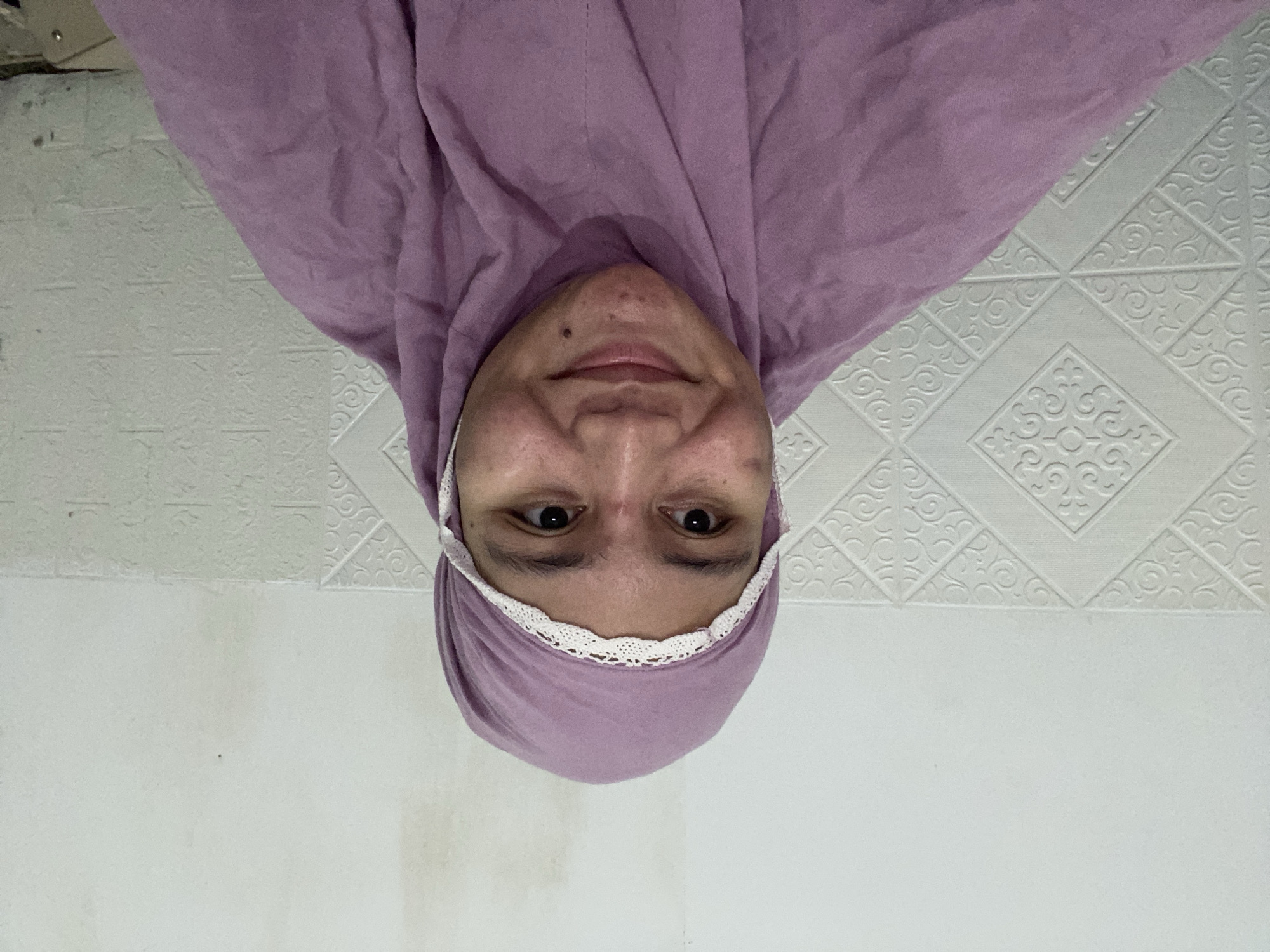 A medium shot shows a person wearing a purple hijab with a white lace trim. The person is looking slightly to their left and smiling. Their face is visible from the forehead to the chin, and they have a mole on their chin. The background consists of a white wall with textured tiles. The lighting appears to be artificial. The image does not contain information about the specific location in Banjarbaru, Indonesia.