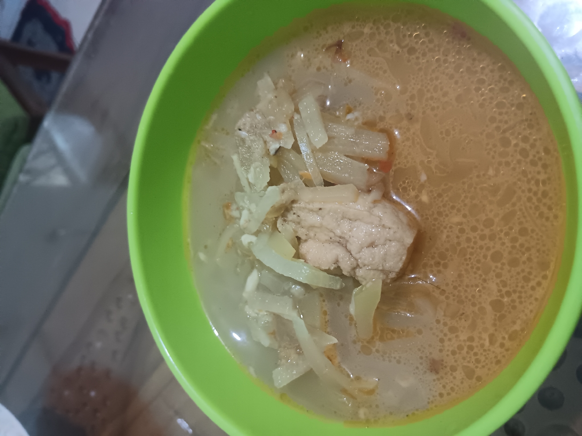 A close-up, high-angle shot shows a bowl of soup. The bowl is bright green. The soup is a light brown color and appears to have small bubbles on the surface. There are pieces of what looks like shredded vegetables and meat in the soup. The soup is served on a surface that appears to be a table made of wood. The background is blurred but suggests an indoor setting. This image was taken in Batursari, Indonesia.
