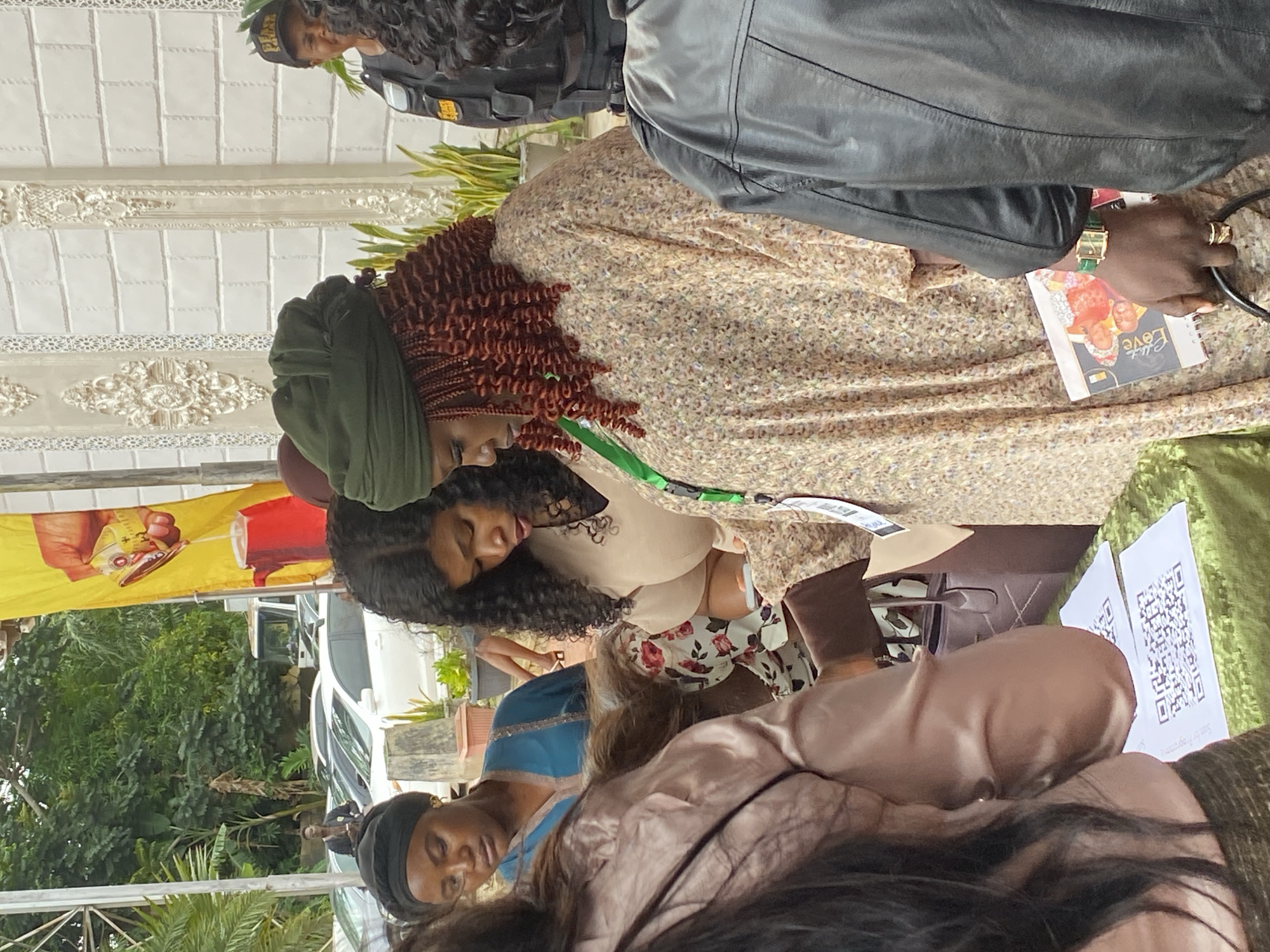 This is a medium shot photograph taken outdoors, possibly in Jos, Nigeria, based on the visual cues. The scene features a group of people gathered around a table covered with a green cloth. On the table, two white papers with QR codes are visible.
In the foreground and midground, several individuals are present. On the left, a woman wearing a dark headscarf and a blue outfit is looking downwards. To her right, another woman with long, curly dark hair is also looking down, seemingly at a mobile device, and wearing a light-colored top. Further to the right and more prominent in the midground is a woman wearing a dark green head wrap and a patterned, loose-fitting dress. Her hair, visible beneath the wrap, appears to be a deep reddish-brown and styled in braids. She is holding a book or magazine with a colorful cover that appears to have images of people on it, and the text "Love" is legible. She is also wearing a green strap across her chest, possibly a lanyard.
In the background, other people are partially visible. A person in a dark tactical vest and a cap is standing on the right side of the frame. Behind the group, there are parked cars, trees, and a building with white brickwork and ornate decorative elements. A large yellow banner with a graphic of a hand holding a measuring tape pouring liquid into a red kettle is also visible in the background. The overall atmosphere suggests an outdoor event or gathering.