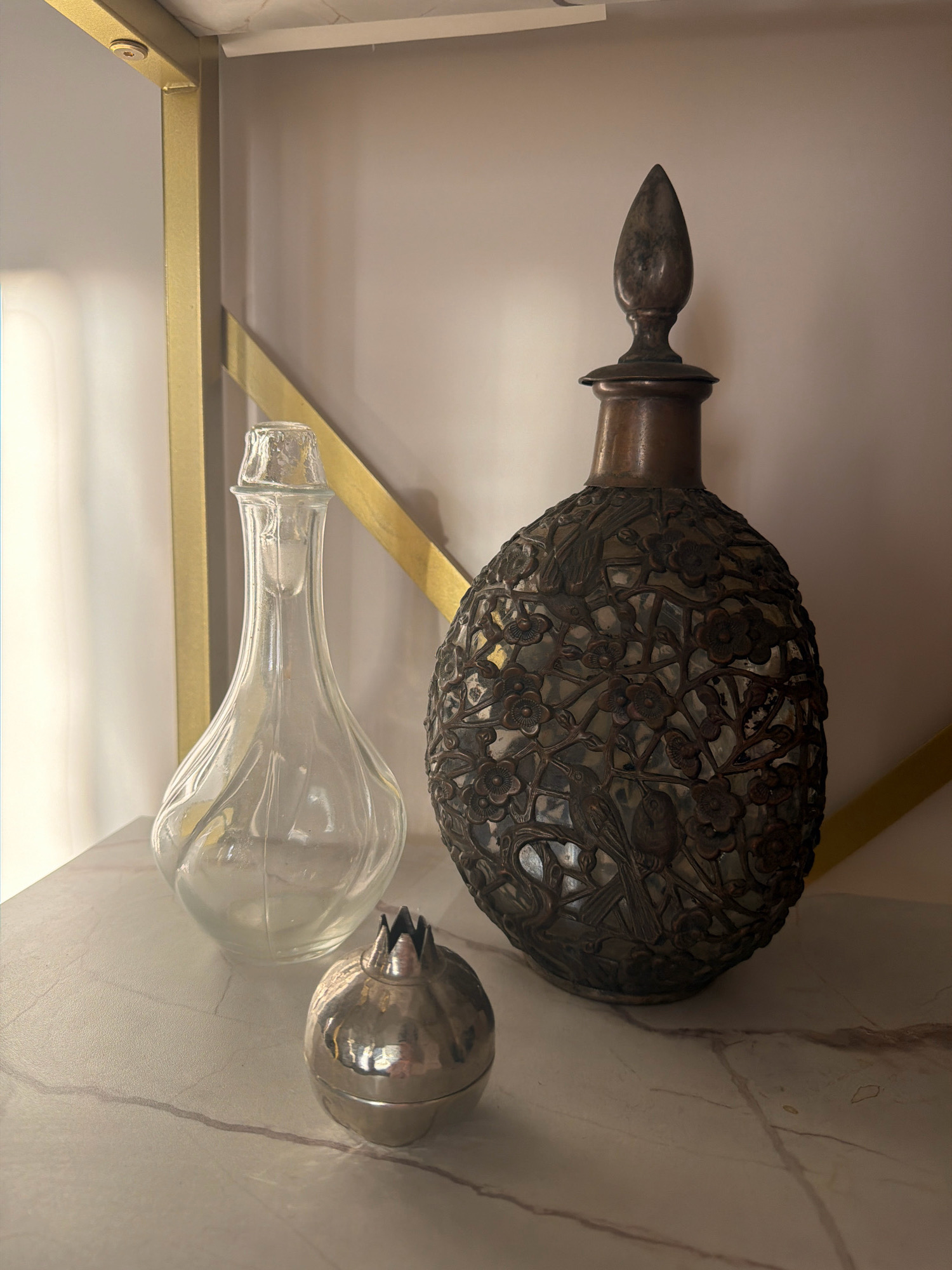 The image shows a still life arrangement on a marble-patterned shelf. The shelf is supported by a gold-colored metal frame. In the foreground, a small, silver, pomegranate-shaped trinket is visible. To its left is a clear glass decanter with a slender neck and a bulbous base. To the right of the decanter is a large, ornate copper-colored bottle covered in intricate metalwork featuring birds and flowers. The background is a plain, light-colored wall. There are no people or any discernible activity occurring in the image. The location is not specified in the image, and therefore, a description of the city of Samara, Russia, cannot be provided.