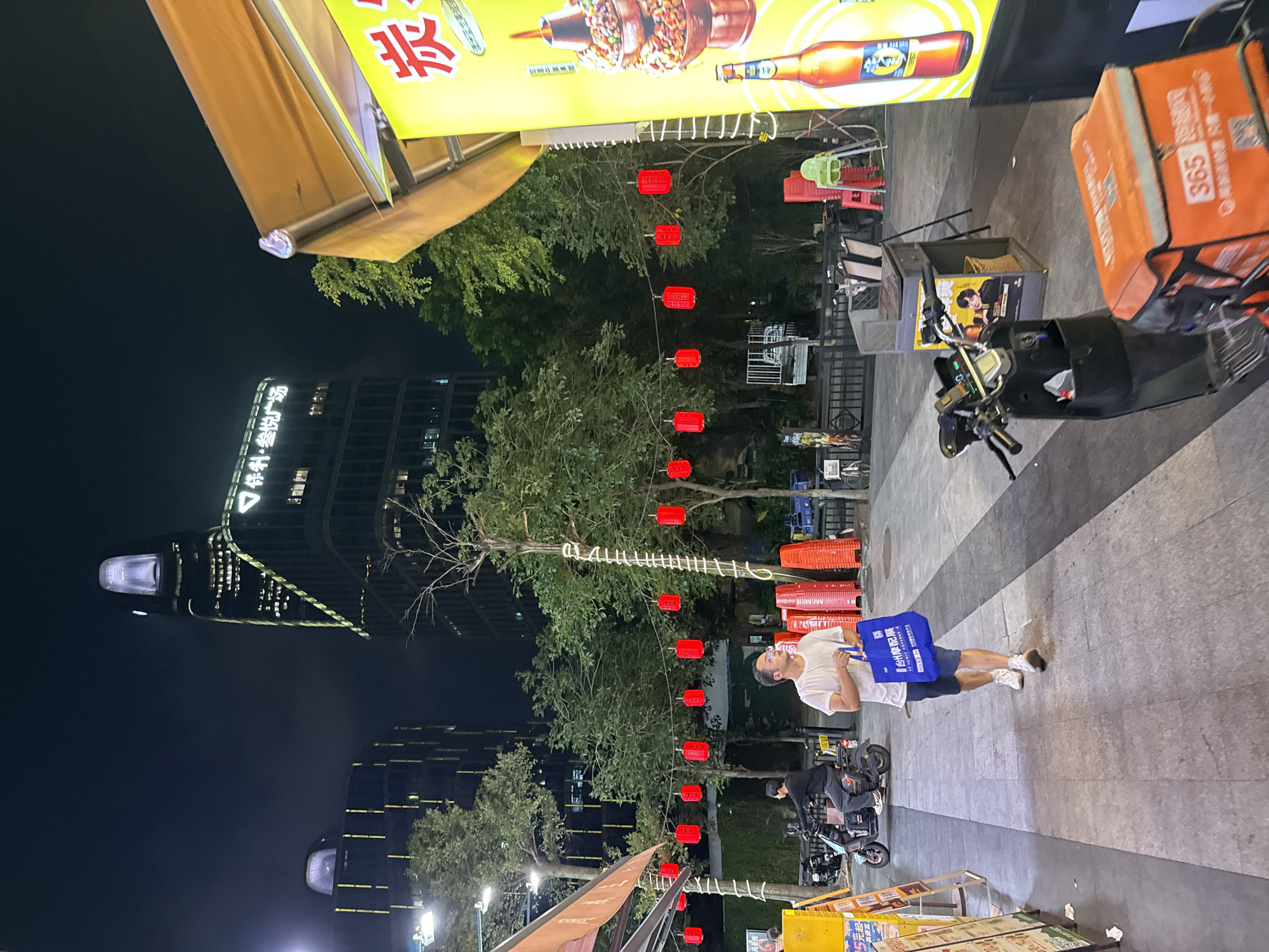 At night, a person wearing a white t-shirt and blue shorts walks on a paved street in Haizhu District, China, carrying a blue shopping bag. He is seen from the waist down, with his face looking to the right and wearing glasses. The street is lined with trees decorated with white string lights and red lanterns. A tall, modern building with illuminated windows and a glowing crown-like structure stands in the background. To the right of the image, there is a food stall with a yellow sign displaying food items and a bottle of beverage. Several electric scooters are parked nearby, one with an orange delivery box. Red chairs and a green high chair are visible near the food stall. In the distance, more trees and buildings are dimly lit.