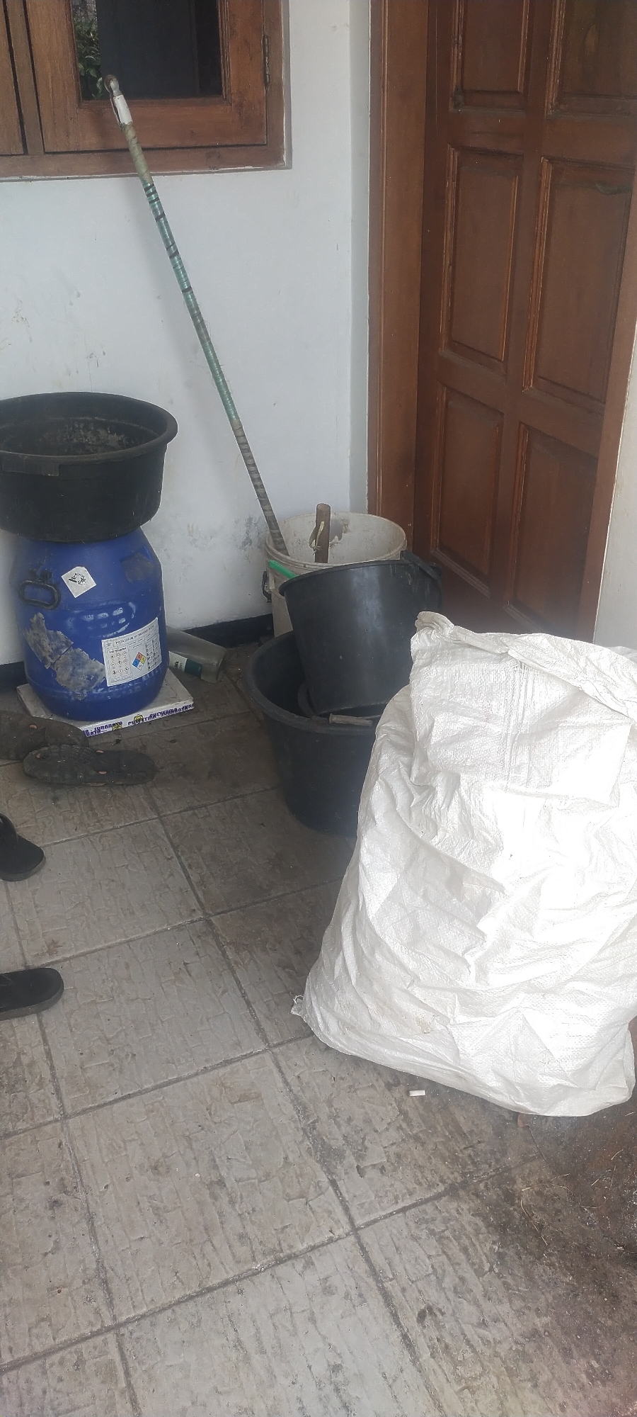 The image depicts an outdoor scene on a tiled floor, likely in the city of Pasirhuni, Indonesia. In the foreground, a large, white woven plastic bag is prominent, filled with an unknown substance and positioned on the right side of the frame. To the left of the bag, several black plastic buckets are stacked or placed on the floor. Behind these buckets, a tall, blue plastic drum with labels is visible, topped with a black basin. A long, green and silver pole, possibly a broom or mop handle, leans against the wall. Further back, a white bucket with a wooden stick protruding from it stands near a wooden door. The wall is painted white, and a wooden window frame is visible in the upper left corner. The floor is made of light-colored square tiles, appearing somewhat grimy. Two black shoe-like objects are partially visible on the floor in the lower left quadrant. The lighting suggests it is daytime, possibly overcast or shaded.
