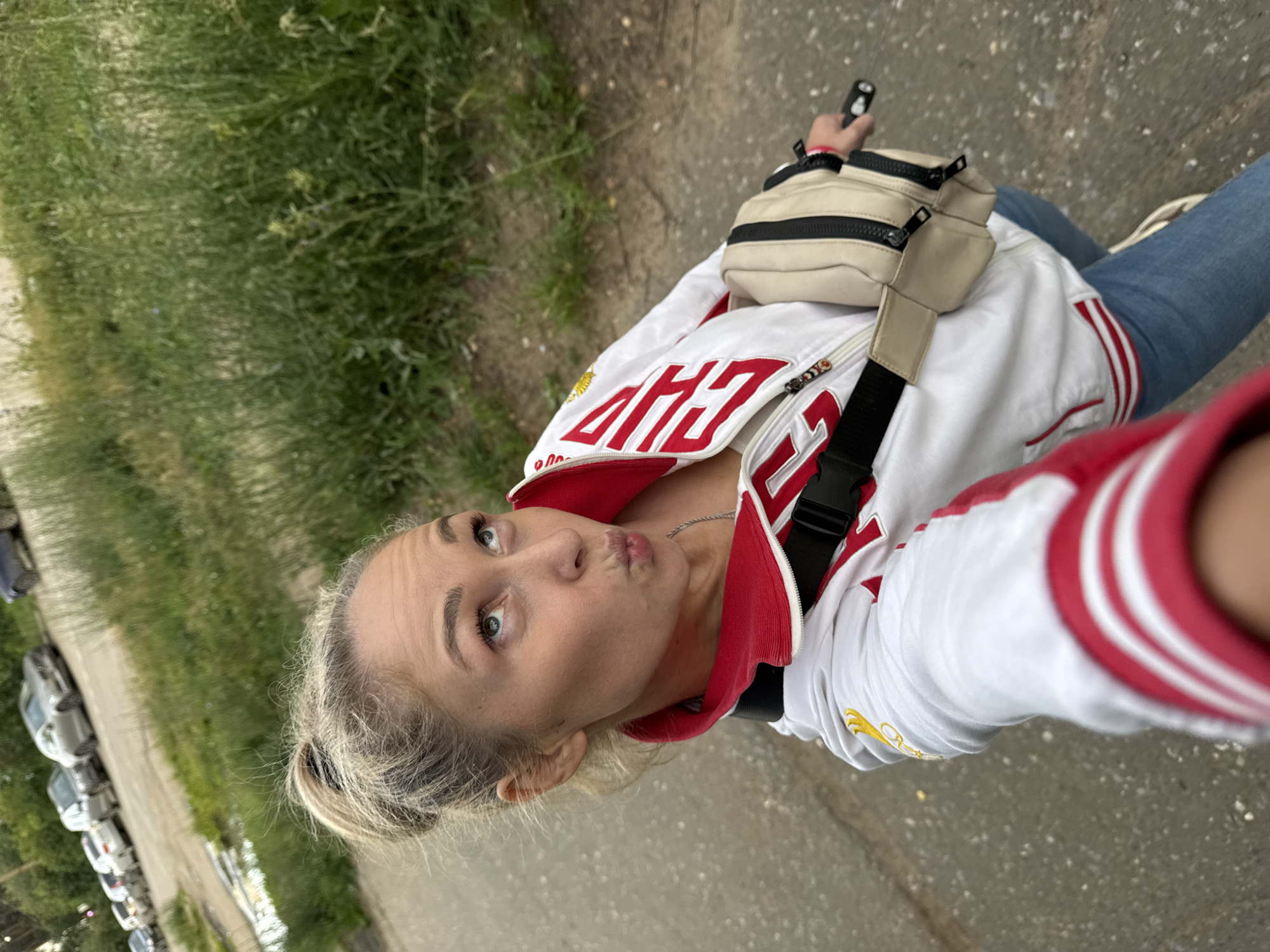 A young blonde woman is taking a selfie outdoors, with a street and grassy area in the background. She is wearing a white jacket with red trim and lettering that says "CHAMP" in red. She also has on blue jeans and a beige fanny pack. The woman is making a "duck face" and looking at the camera. There are cars parked along the street in the background, and the weather appears to be overcast. The scene is likely in Chaikovsky, Russia, as indicated by the location context. The woman appears to be in a casual, relaxed mood, possibly on a walk or outing. The image is taken from a high angle, looking down at the woman.