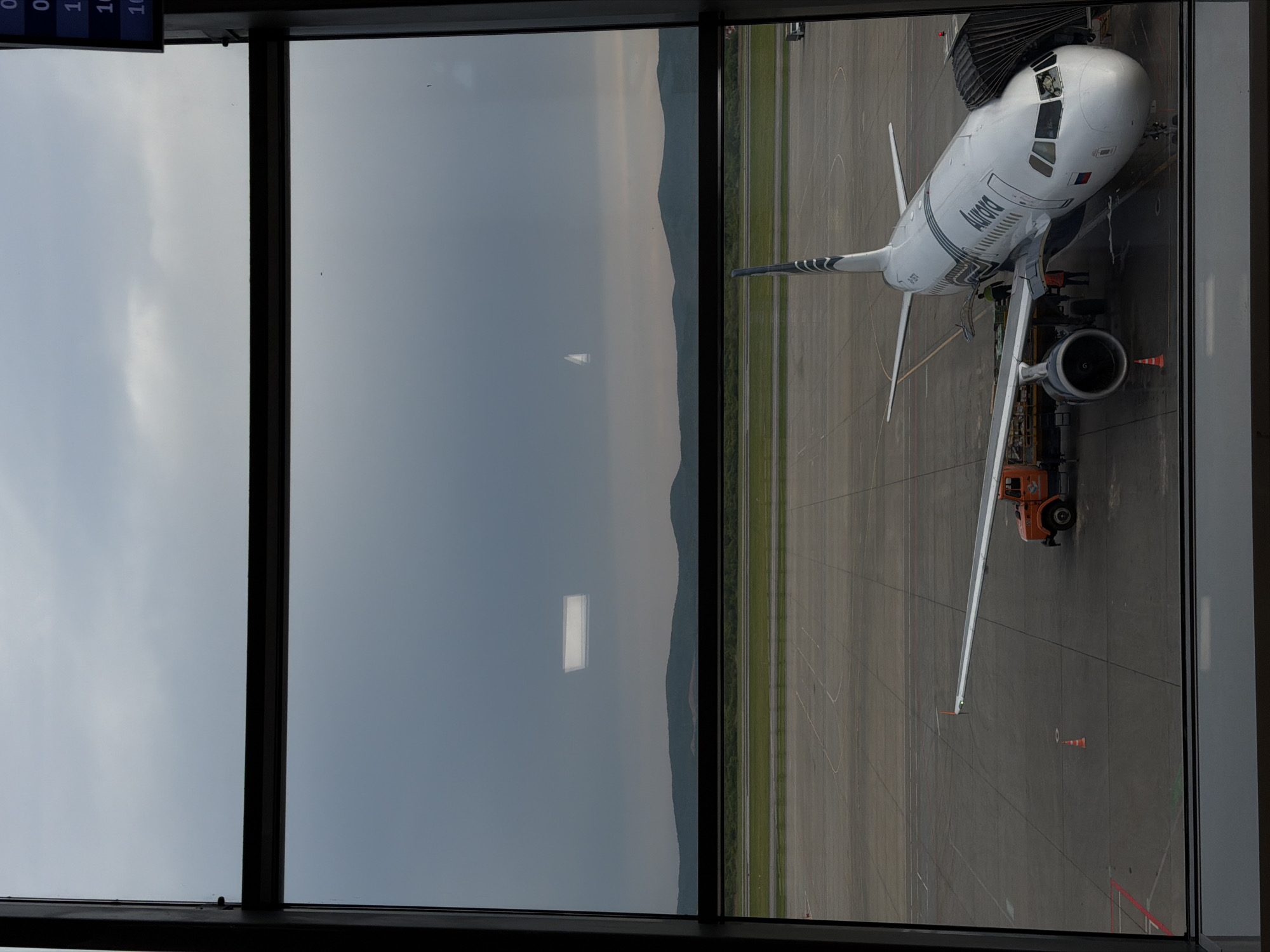 A view from a window looking onto the tarmac at Artyom Airport in Russia. An "Aurora" airlines plane is docked to a terminal via a jet bridge. The plane has a utility vehicle under its wing. Beyond the tarmac, a grassy field borders an undefined mountainous area. The sky is overcast.