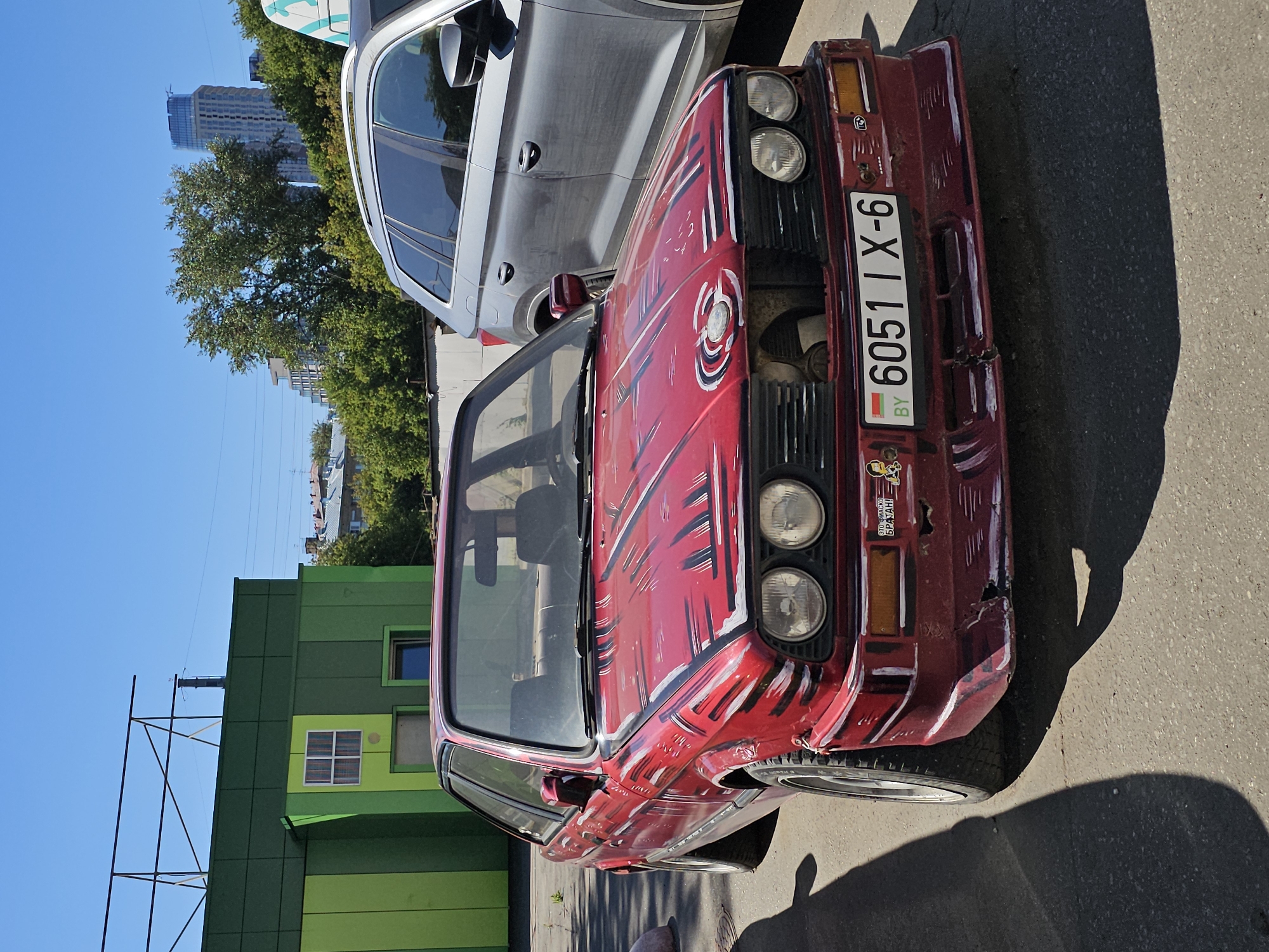 The image shows a red BMW E30 coupe parked on an asphalt surface, possibly a parking lot. The car has a custom paint job on the hood, featuring white and black stripes. A Belarusian license plate, reading "6051 IX-6", is visible on the front of the car. To the right of the BMW, a silver SUV is parked. In the background, a modern building with a glass facade and a green building with striped panels are visible. Trees and a clear blue sky complete the scene. The context of Moscow, Russia, is not directly inferable from the visual elements within the image itself.