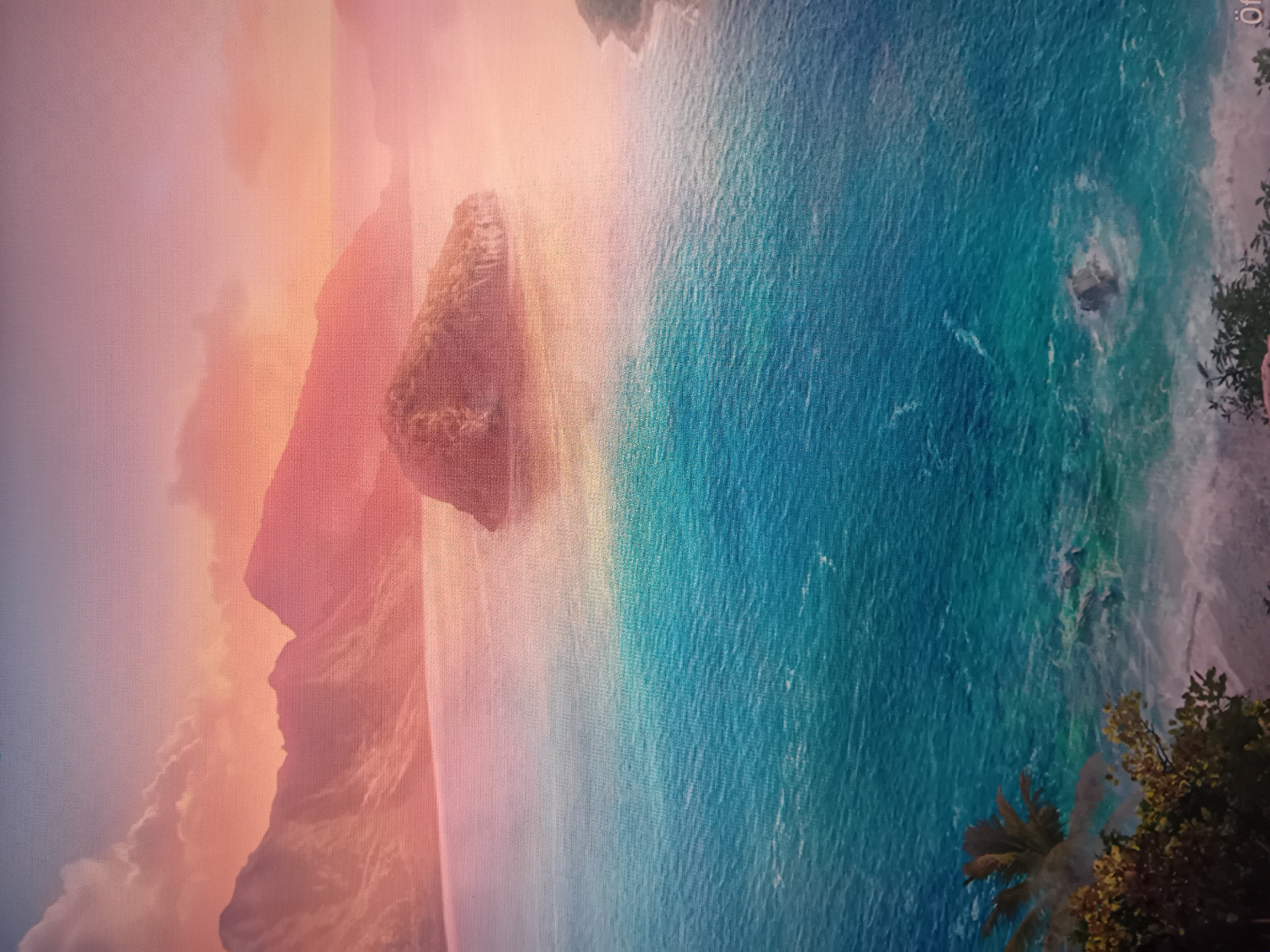 A vibrant tropical coastal landscape is depicted at either sunrise or sunset. The image displays a clear grid-like or pixelated texture, suggesting it is a photograph of a screen.
The sky on the left and upper-middle is awash with soft, warm hues of pink, orange, and light purple, hinting at either dawn or dusk. Dominating the left side are tall, rugged mountains or cliffs, largely silhouetted against the colorful sky, with some textural detail visible on their surfaces.
An expansive body of water, likely an ocean or large bay, stretches across the middle and right of the frame. The water transitions from lighter, pastel tones near the horizon to rich shades of turquoise and deep blue closer to the foreground, with visible ripples and waves on its surface. In the mid-distance within the water, a distinctive rocky island or formation stands prominently.
The right side of the image features a sandy shoreline, although the sand itself is mostly out of view. Lush, green tropical foliage, including clear palm fronds and other leafy trees, frames the far right edge of the scene, indicating a verdant coast.
No specific activity or event is taking place; it is a serene landscape. The time of day is clearly indicated by the soft, warm sky colors. The location cues point to a tropical environment. In the top-right corner of the image (when oriented correctly), a faint, small symbol resembling "O:" is visible.