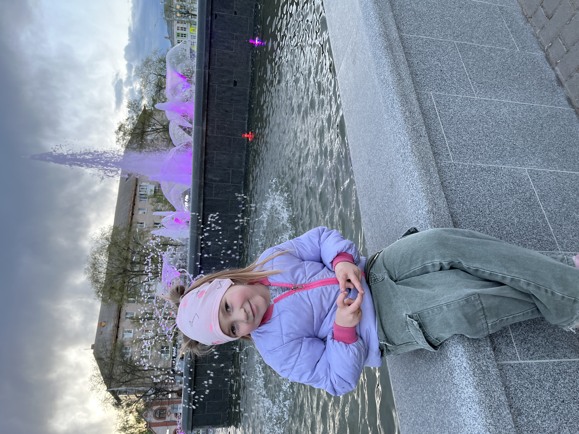 A young girl is sitting on the edge of a fountain in the city of Chaikovsky, Russia. She is wearing a light purple jacket, pink and gray undershirt, and green pants. Her hair is blond and she is wearing a pink headband. The fountain has purple lights and water spraying in various directions. Buildings can be seen in the background. The sky is cloudy.
