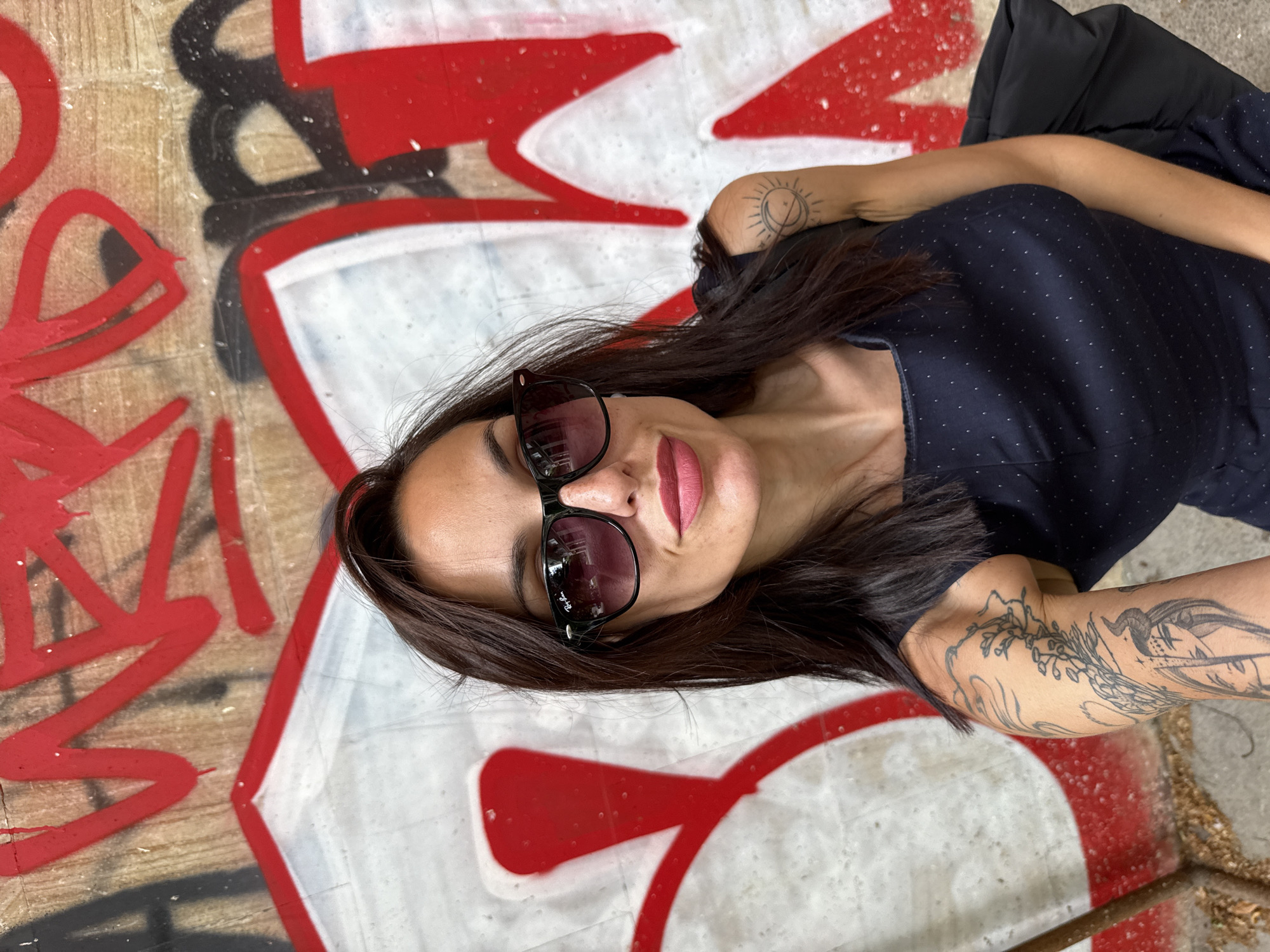 A woman wearing sunglasses and a dark blue dress with white polka dots lies on a surface with graffiti, consisting of red and white paint, with some black elements. The location is suggested to be in the city of Pleven, Bulgaria. The woman has dark hair and tattoos on her arms. A black jacket is visible next to her.