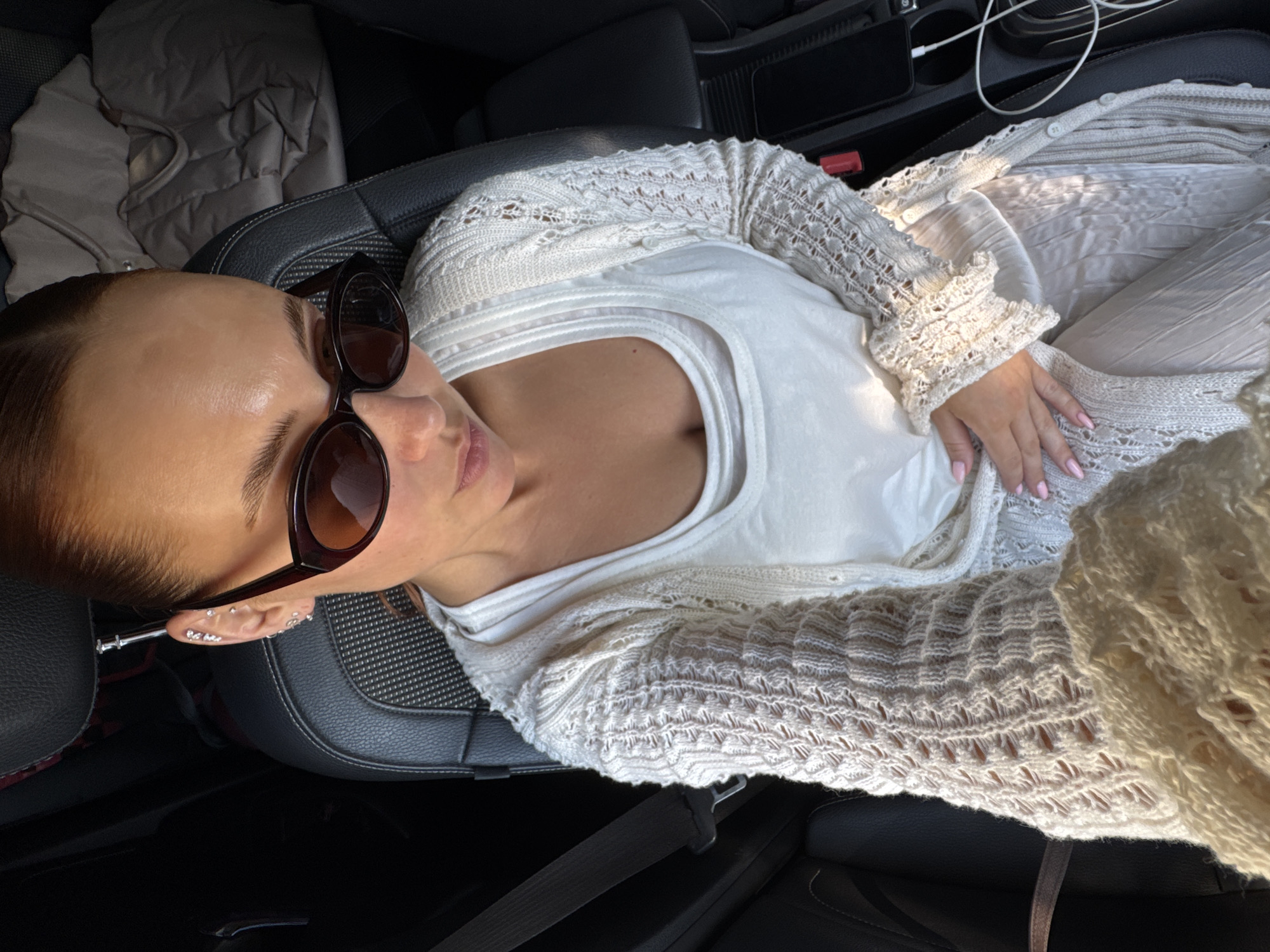 A woman lies in the passenger seat of a car. She wears a white tank top, a white cardigan, and sunglasses. Her hair is pulled back, and she has multiple piercings in her left ear. The car interior is visible, including the black leather seats, center console, and a jacket. The image was not taken in Chaikovsky, Russia.