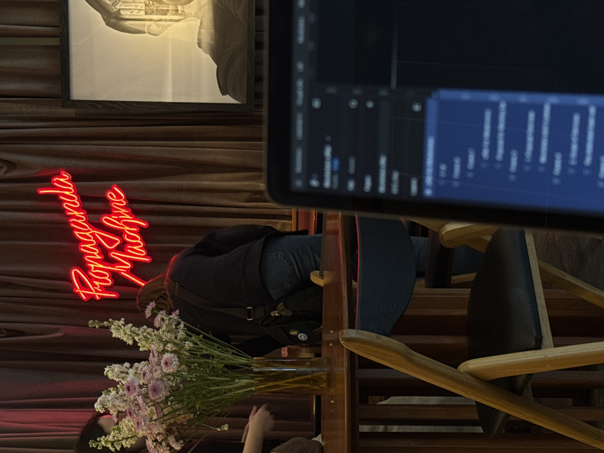 This image captures an intimate indoor scene, likely a modern cafe, bar, or creative workspace in Yerevan, Armenia, during the evening. The setting is characterized by dark wooden paneled walls and sophisticated wooden furniture.
A prominent feature on the left is a vibrant red neon sign, oriented vertically, displaying the words "Propaganda Machine" in a flowing script, casting a warm glow on the surroundings. Above the neon sign, a framed monochromatic artwork, possibly depicting a classical sculpture or architecture, hangs on the wall.
In the foreground, a person is seated at a wooden table, their back partially facing the viewer. They are wearing a dark top, blue jeans, and have a dark bag adorned with various pins. Their posture suggests they are engaged in an activity at the table. A large, lush bouquet of white and light purple flowers in a clear glass vase sits on the table, partially obscuring the seated individual. The hand of a second person is visible on the lower left, near the flowers, indicating another individual's presence at the table.
To the right, a large monitor screen is partially visible and slightly out of focus. It displays a dark user interface with numerous blue and white graphs and bar charts, suggesting the use of software for data analysis, audio visualization, or video editing. The overall lighting is dim, creating a cozy and focused atmosphere, with the neon sign providing a key light source. Wooden chairs with dark cushions are also visible, completing the interior decor.