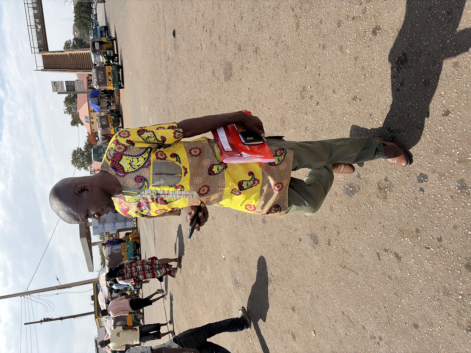 The image captures a vibrant street scene in Jos, Nigeria, under bright daylight, evidenced by prominent shadows. The primary subject is a man in the foreground, positioned slightly to the left, wearing a distinctive yellow shirt with brown and red patterns, green trousers, and brown shoes. He is looking down at a smartphone in his left hand, while cradling a red package and a black diary, clearly marked "2026," in his right arm.
Behind the main subject, the street is bustling with activity. Several individuals walk along the paved road. A line of colorful tricycles, known as Keke Napep, are parked or moving slowly. Some of these vehicles display visible identification numbers such as "PTA-10529" and "PTA 7224".
In the background, an elevated pedestrian bridge or walkway stretches across the scene. On the right side of the street, a petrol station is visible, featuring a "TotalEnergies" sign with numerical values "1305", "1990", and "220" – likely indicating fuel prices or types – and the word "QUARTZ". Other partial text elements like "ANGT" and "GTATINGT" are also visible on structures in the distance. The sky is clear, suggesting fair weather. The scene depicts a typical, busy urban environment.