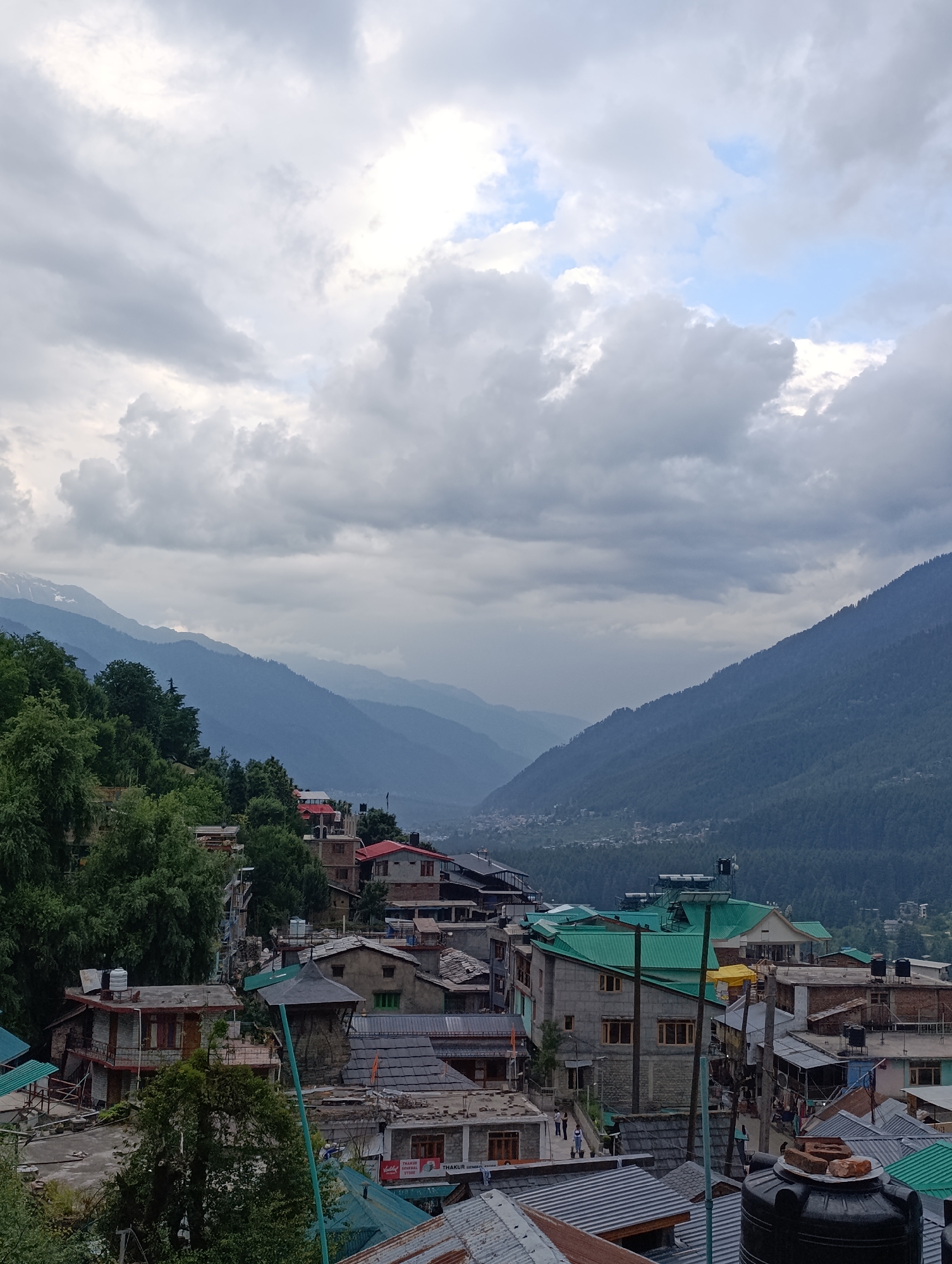 This media file depicts a cloudy day in Vashisht, India, a village known for its hot springs. The scene is a sprawling hillside village nestled in a valley, with layers of misty mountains receding into the distance. The architecture is dense, with buildings constructed of stone and brick, many featuring brightly colored green roofs. Homes are clustered together, with narrow pathways and alleys winding between them.
In the foreground, we see several buildings with a mix of modern and traditional elements. Some have visible water tanks on their roofs, suggesting a reliance on collected water. There are hints of everyday life, with a few people visible walking along the paths and near the buildings. A sign for "THAKUR GENERAL STORE" is visible on one of the buildings.
The weather is overcast, with dramatic, thick clouds filling the sky. While the sun is not directly visible, the light is diffused, creating a soft, somewhat somber atmosphere. The mountains are shrouded in a bluish haze, indicating distance and possibly humidity. The overall impression is one of a lived-in, rural community set against a breathtaking natural landscape, with the weather adding a touch of dramatic intensity to the scene. The time of day appears to be late morning or early afternoon, given the amount of ambient light despite the cloud cover.