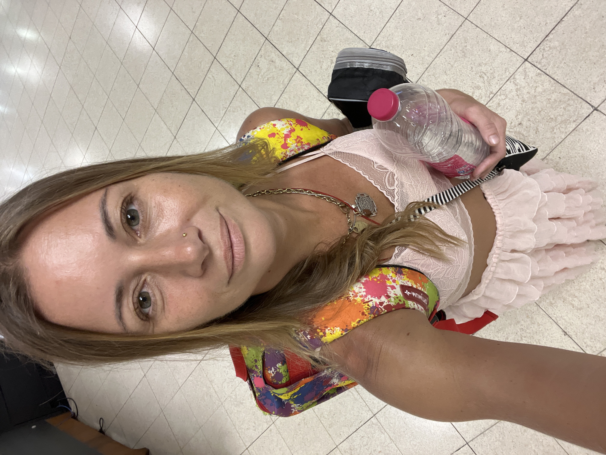 A woman, identified as being in Katunayake, Sri Lanka, is captured in a selfie. She has light skin, green eyes, and shoulder-length blonde hair. She is wearing a pink lace bralette, a frilly pink skirt, and a backpack with a colorful, paint-splatter design. Around her neck, she has a silver chain with multiple pendants. In her right hand, she is holding a clear plastic water bottle with a pink cap. She is standing on a tiled floor. The background includes what appears to be a dark object on the left side of the image, possibly furniture, and the tiled floor continues to the right.