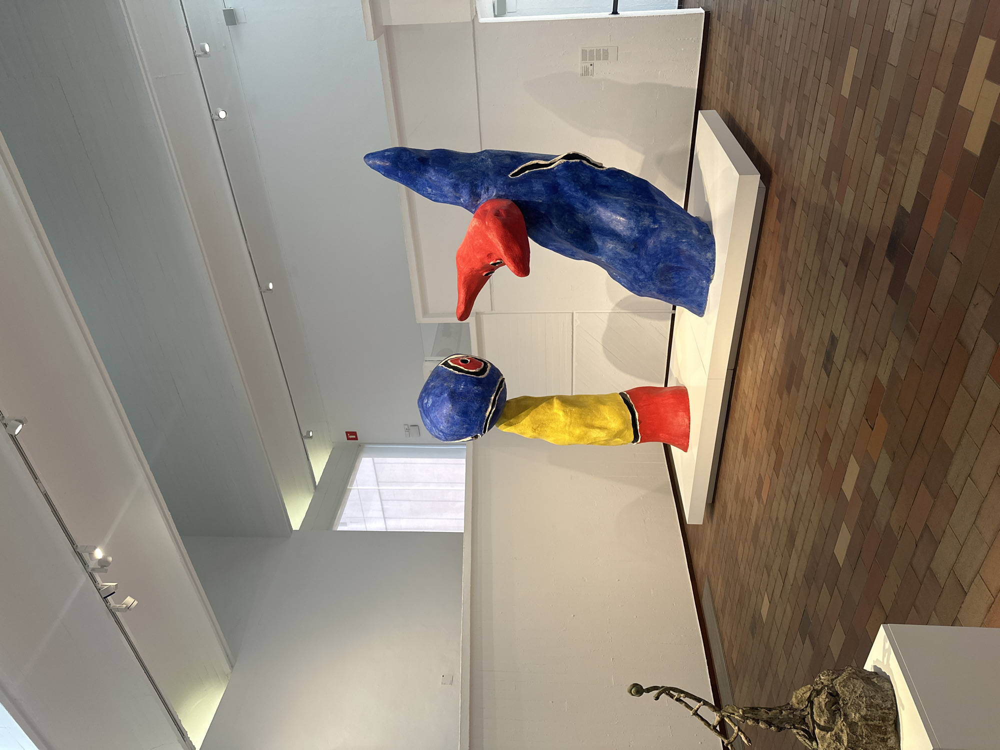 The image captures a colorful modern art sculpture, specifically Joan Miró's "Dona i Ocell" (Woman and Bird), in the Parc de Joan Miró in Barcelona, Spain. The sculpture, predominantly blue, red, and yellow, stands on a white plinth. The indoor setting is an art gallery with a high ceiling featuring track lighting and white walls. The floor is covered with brick tiles. Another sculpture, partly visible in the bottom right, is also placed on a white plinth.