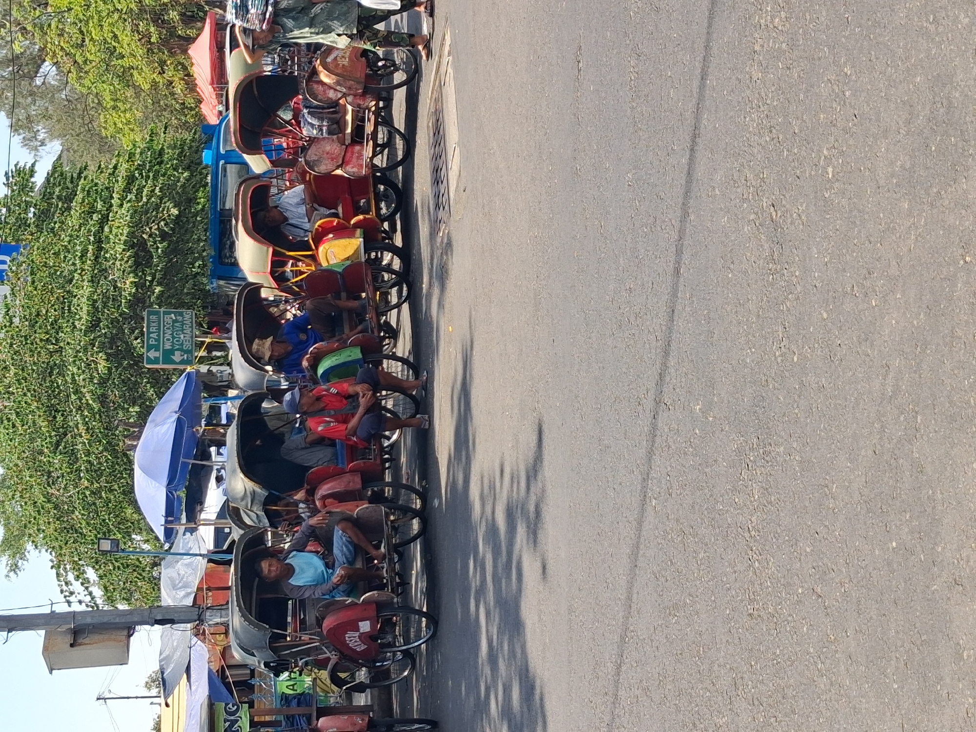 The image captures a street scene in Surakarta, Indonesia, likely near a tourist area or transportation hub. A row of becaks, a type of three-wheeled passenger vehicle, is parked along the side of a paved road. Several becak drivers are seated in their vehicles, some appearing to be waiting for customers. One driver in a blue shirt is in the foreground, while others in red and casual attire are further back. A person in a camouflage pattern shirt and dark pants walks past the becaks. Overhead, a large green tree canopy shades a portion of the scene, and a blue and white umbrella is visible above the becaks. A directional sign with text in Indonesian, indicating "PARKIR WONOGIRI YOGYA SEMARANG" (Parking Wonogiri Yogyakarta Semarang), is mounted on a pole. Tarpaulins and what appear to be makeshift stalls are set up behind the parked becaks, suggesting a market or vendor area. The overall atmosphere is that of a busy, daytime street in a tropical urban environment.