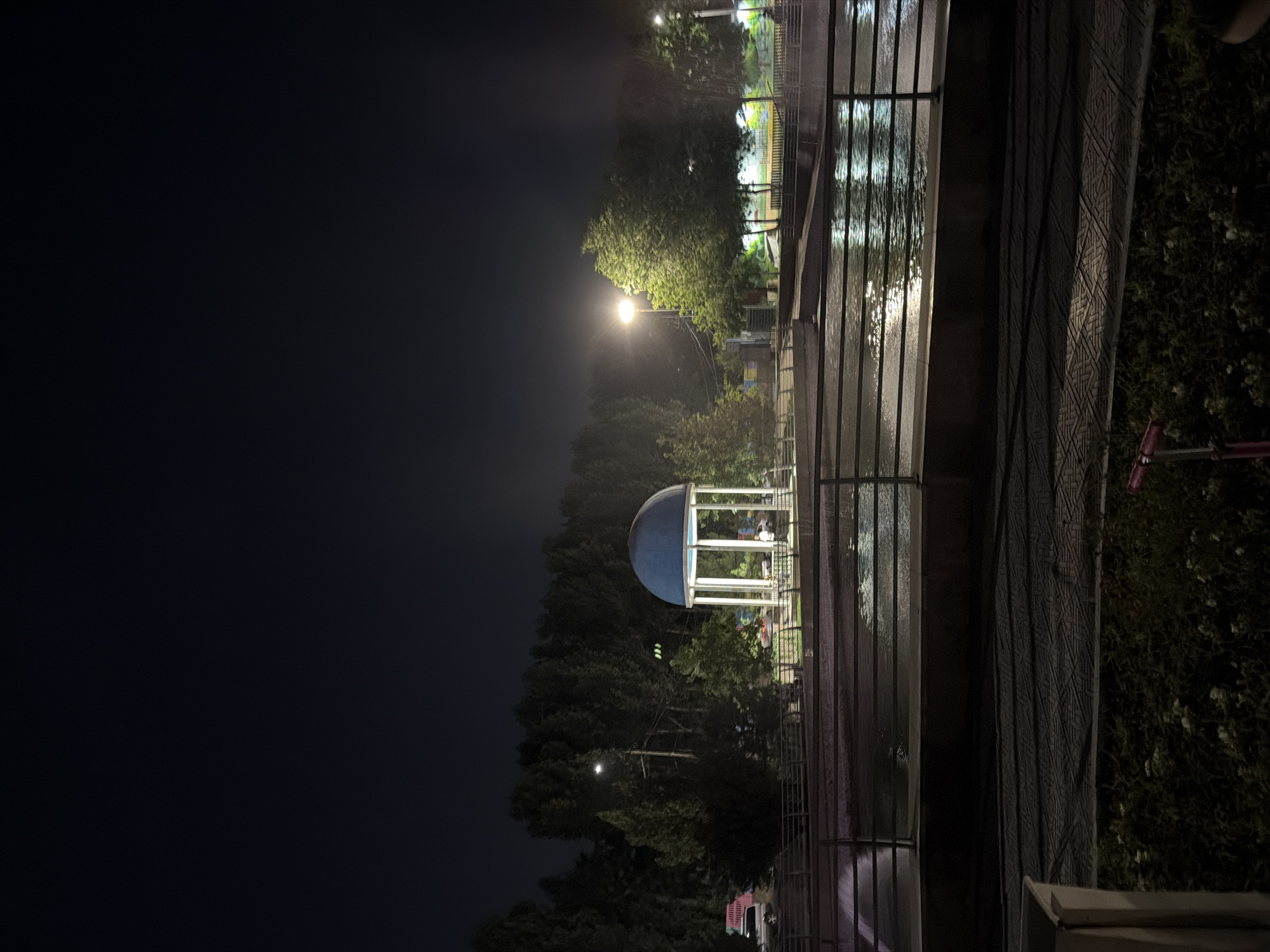This media file depicts a night scene in Shymkent, Kazakhstan. The image is oriented vertically, showing a park-like setting illuminated by artificial lights.
The primary subjects include a blue-domed gazebo with white pillars, situated amongst lush green trees. Several figures are visible inside and around the gazebo, suggesting a gathering or event is taking place. A street lamp with a bright, warm light illuminates a portion of the trees and the path in front of the gazebo.
To the right of the gazebo, a paved area with water features or reflections is visible, bordered by a metal railing. This area is also lit, creating shimmering patterns of light on the wet surface. Further to the right, dense foliage and a patterned wall or covering are visible, with a hint of red object, possibly a scooter, in the background. The overall ambiance is one of a public space at night, potentially a park or recreational area.
The time of day is clearly night, due to the darkness of the sky and the reliance on artificial lighting. The weather appears to be clear, as indicated by the reflections on the wet surfaces, which could be due to recent rain or a water feature. There are no overt indications of any specific event, beyond the presence of people in the gazebo, which could be a casual meeting or a more planned occasion. The scene evokes a sense of calm and perhaps a bit of mystery due to the night lighting. No visible text is present in the image.