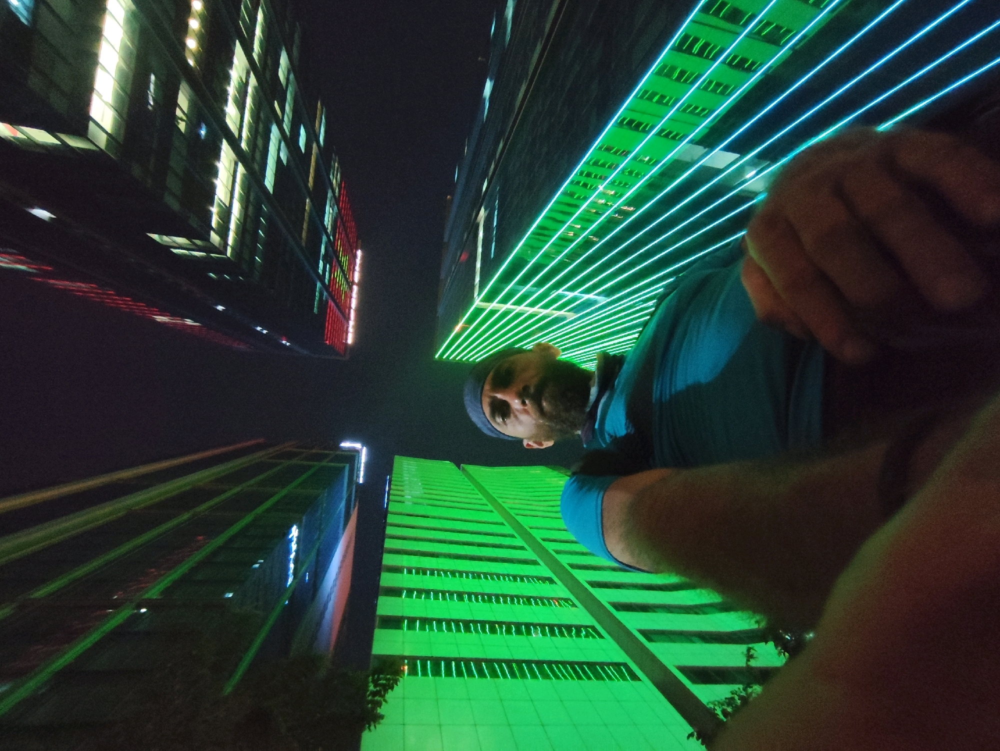 A man, viewed from a low angle, appears to be taking a selfie in front of illuminated skyscrapers at night in Ho Chi Minh City, Vietnam. The man is wearing a blue t-shirt and a dark blue beanie. His right hand is visible, obscuring part of his face and the camera. The background is dominated by modern buildings with extensive glass facades, reflecting the dark night sky and the vibrant lights of the city. One building on the right is covered in a bright green neon light display that runs vertically in parallel lines. Another building on the left has horizontal rows of yellow and white lights. A third building further left displays red vertical lights. The perspective suggests the photo was taken from street level or a lower floor of a building. The overall scene captures the dynamic and brightly lit urban landscape of Ho Chi Minh City at night.