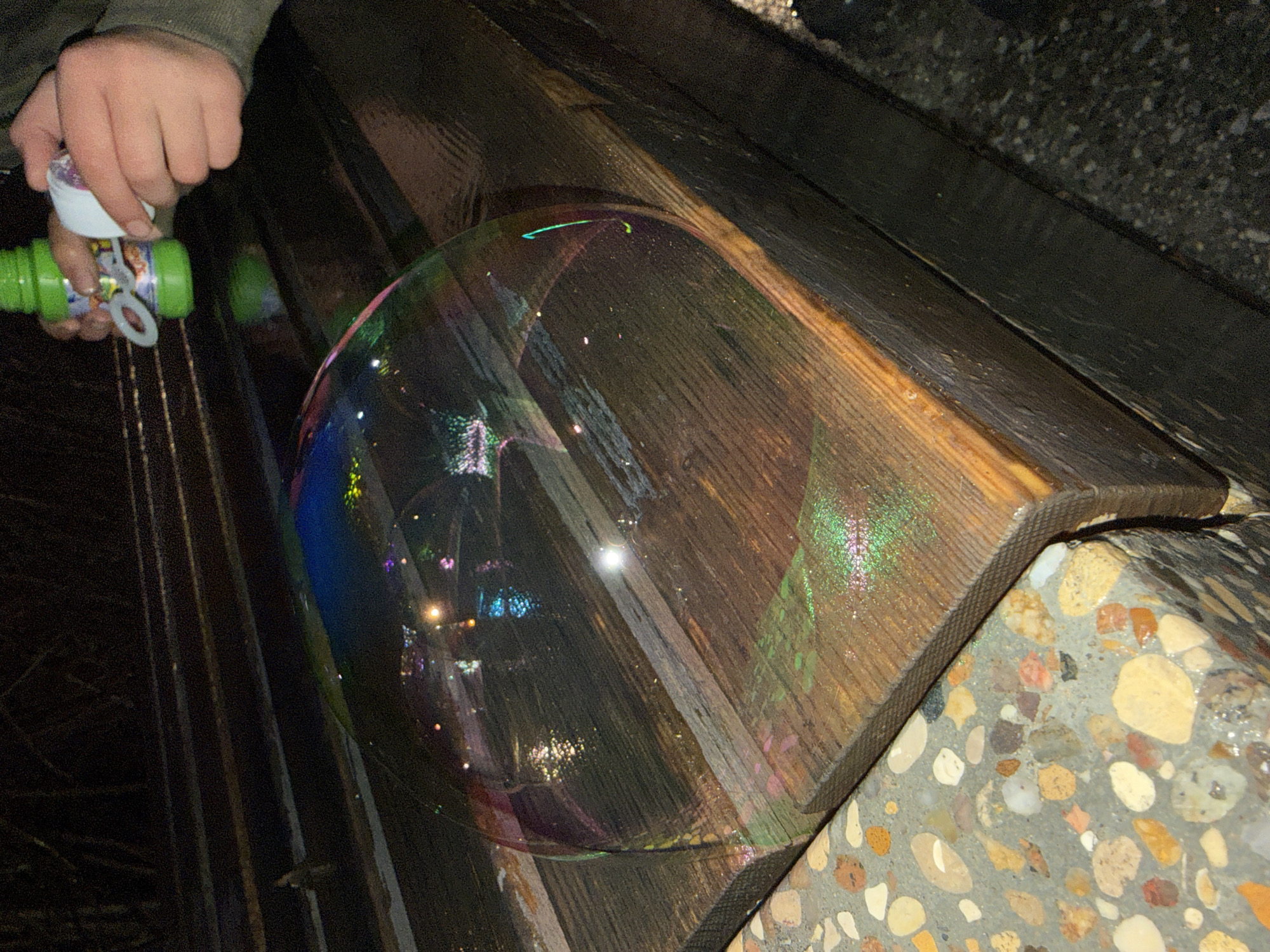 A person's hand holds a green bubble wand bottle near a large bubble resting on a wooden bench. The bench sits on a concrete surface with embedded pebbles. The bubble reflects surrounding lights. This scene is located in Dmitrov, Russia.