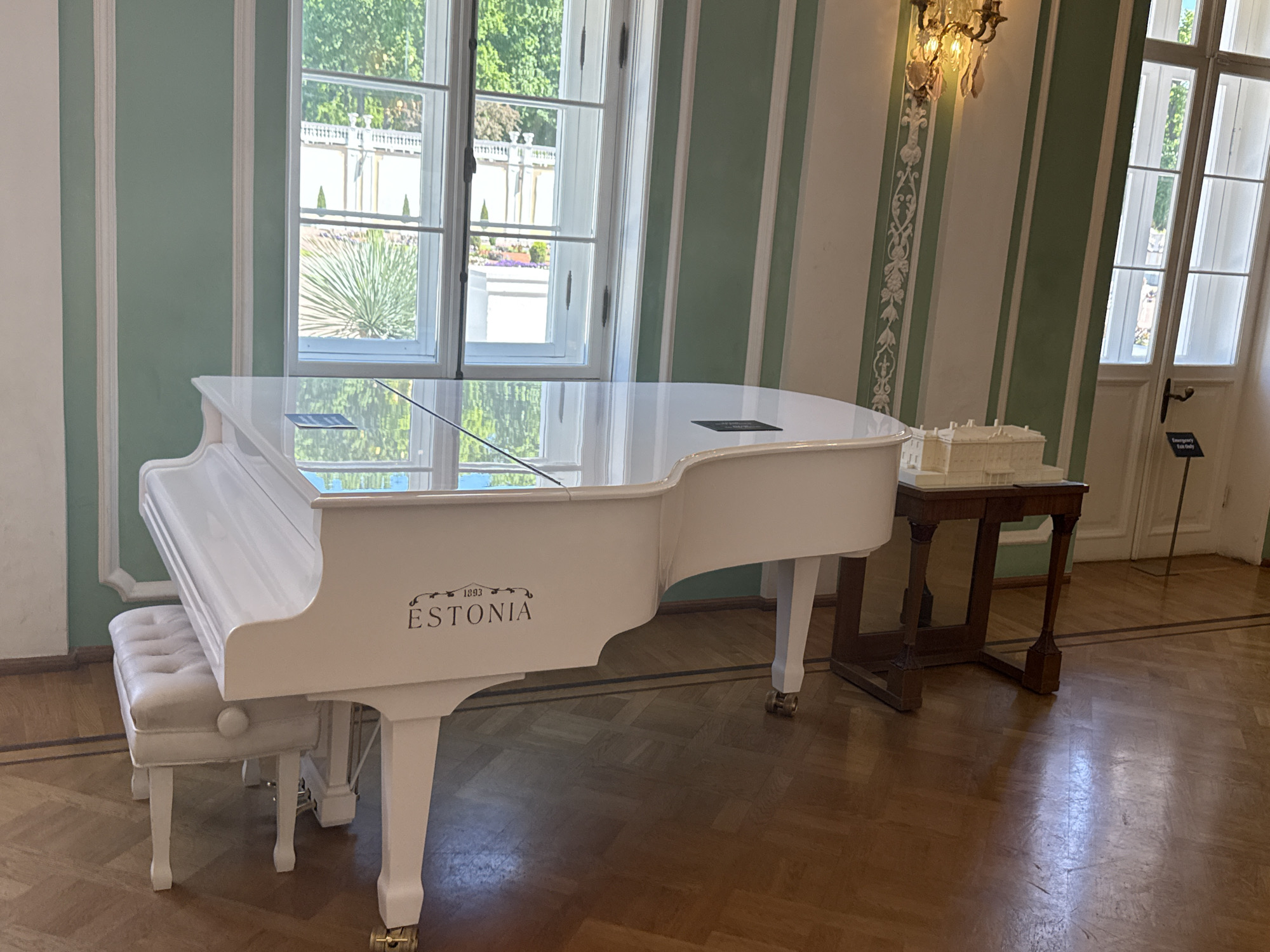 A white Estonia grand piano and matching upholstered bench sit on a polished wooden floor in a room with pale green and white striped walls and large windows. A small, detailed architectural model of a building rests on a dark wooden table to the right of the piano. The windows look out onto a green garden with trees and formal plantings. A chandelier hangs from the ceiling, and an "Emergency Use Only" sign is visible on a door in the background. The scene is well-lit by natural light and artificial lighting from the chandelier. There are no people in the image.