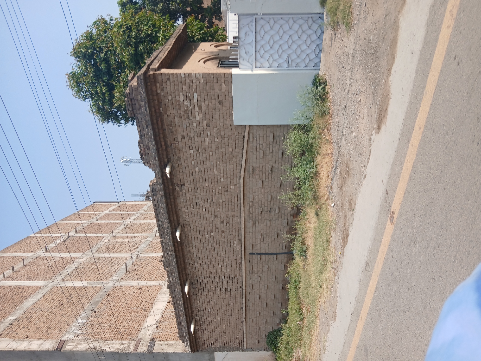 In Peshawar City Tehsil, Pakistan, a low-rise brick building with a tiled roof stands adjacent to a much larger, unfinished concrete and brick structure. The smaller building features several lights mounted on its facade and a decorative metal gate. The area is overgrown with grass and other vegetation. Overhead, power lines crisscross the clear blue sky. The scene is captured from a street-level perspective, suggesting a public thoroughfare adjacent to the properties.