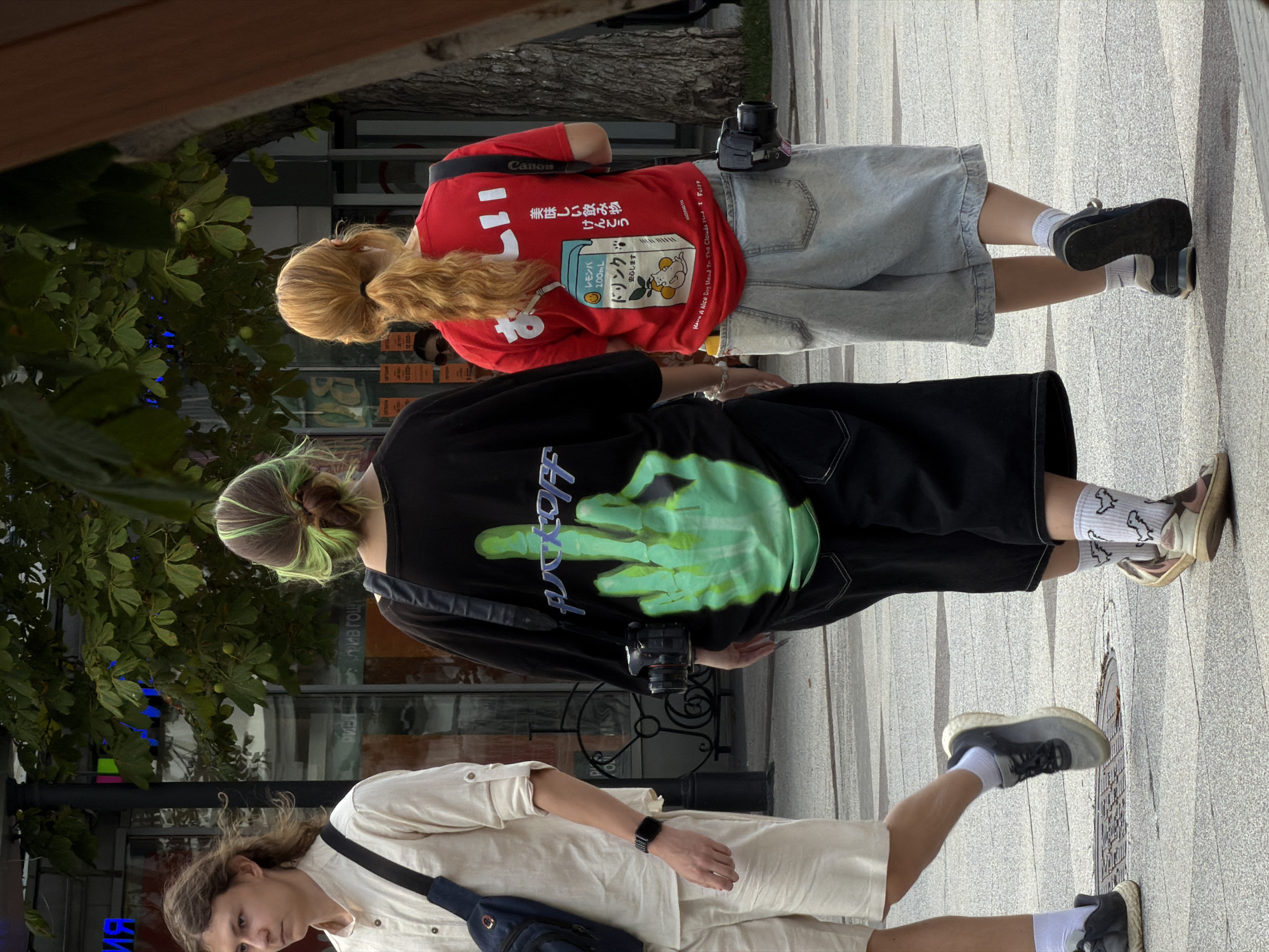 The image captures a street scene with three individuals walking outdoors. On the left, a person with medium-length wavy brown hair and wearing a light-colored collared shirt and light-colored shorts walks away from the camera. They have a dark blue bag slung across their chest and a dark smartwatch on their left wrist.
In the center, a person with their back to the camera has dark hair with streaks of bright green and is wearing a black t-shirt featuring a large, neon green skeleton hand giving the middle finger and the word "FUCKOFF" above it. They are also wearing black shorts. A black camera is attached to a strap around their neck, with the camera body hanging at their side.
To the right, a person with auburn hair tied in a ponytail is walking away from the camera. They are wearing a bright red t-shirt with Japanese text and graphics of a milk carton and an anthropomorphic cat. They are also wearing light-wash denim shorts. A black Canon camera is attached to a strap around their neck, with the camera body hanging at their side. White earbuds are visible in their ears.
The background shows a street with trees, storefronts with signage, and streetlights. The ground is paved with textured concrete. The overall impression is of a casual urban environment. This scene is consistent with a location in Saratov, Russia, based on the Cyrillic text visible in the background signage.