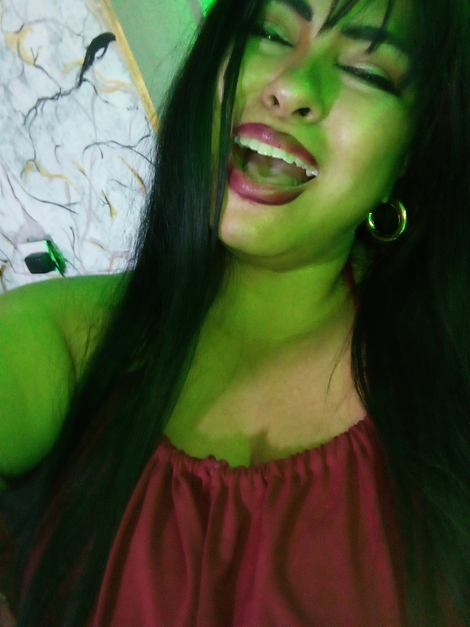 A woman with dark hair and green-toned skin is shown from a low angle, wearing a maroon halter top and gold hoop earrings. She has a wide, open-mouthed expression. The background is indistinct, with a pattern of black lines and shapes on a white surface partially visible. The location is unknown.