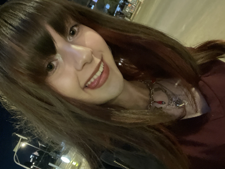 A smiling woman with brown hair and bangs poses for a selfie. Her face is tilted slightly towards the viewer. She has light skin and her lips are slightly parted, revealing her teeth. She wears a necklace with a red fish charm and a Star of David pendant. The background is blurry, with what appears to be streetlights and buildings visible in the distance. The lighting suggests it is evening or night.