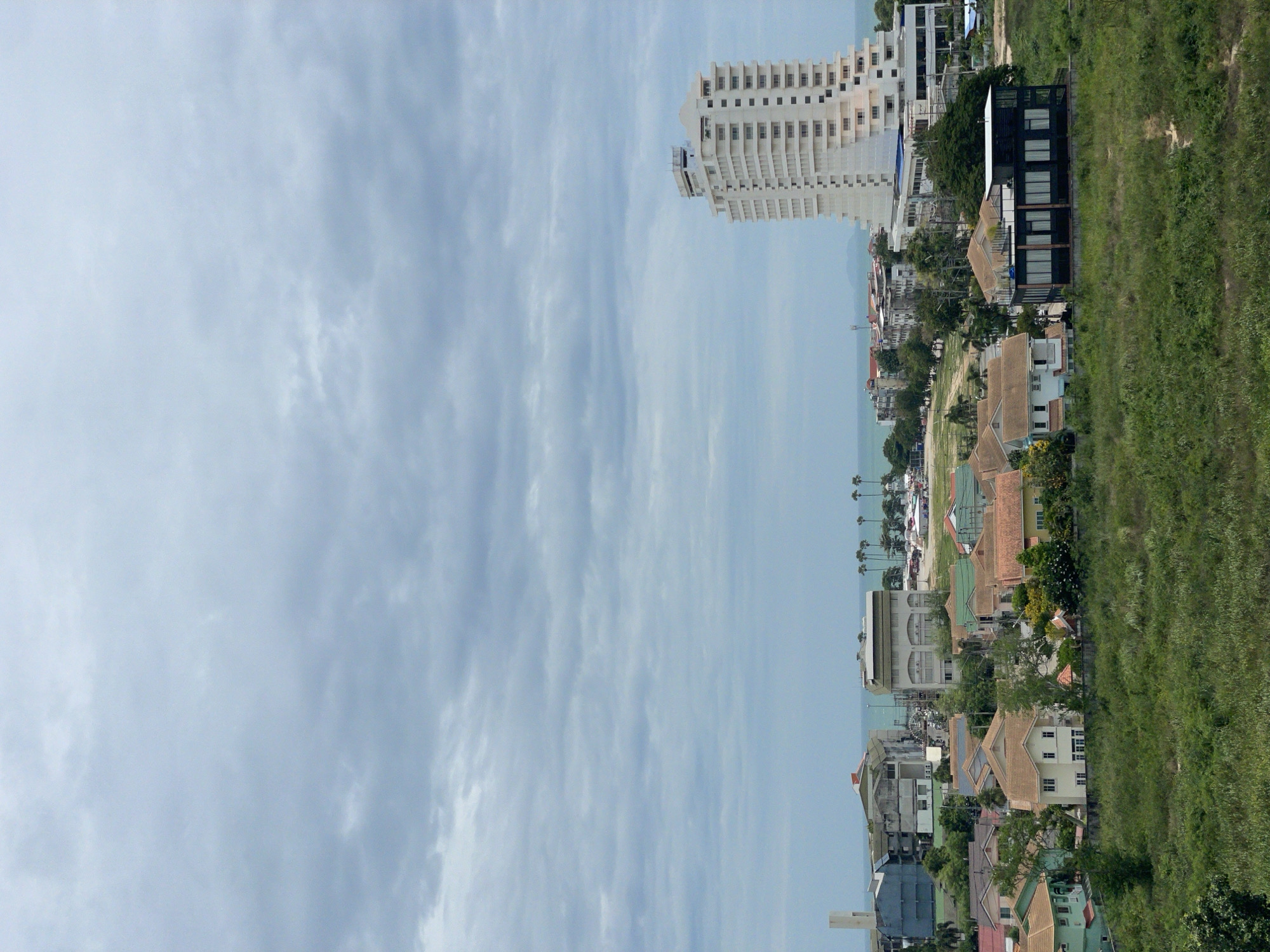 The image captures a panoramic view of a coastal city, likely Pattaya City, Thailand, on a cloudy day. The foreground is dominated by lush, green vegetation. In the middle ground, a cluster of residential buildings with red and brown tiled roofs is visible. Beyond these homes, a modern, multi-story white building with a distinctive stepped design stands prominently, suggesting a hotel or condominium complex. Further in the distance, the tranquil sea stretches out, meeting the hazy horizon. Scattered palm trees dot the landscape, indicating a tropical climate. The sky is overcast with thick, grey clouds, creating a subdued lighting condition.