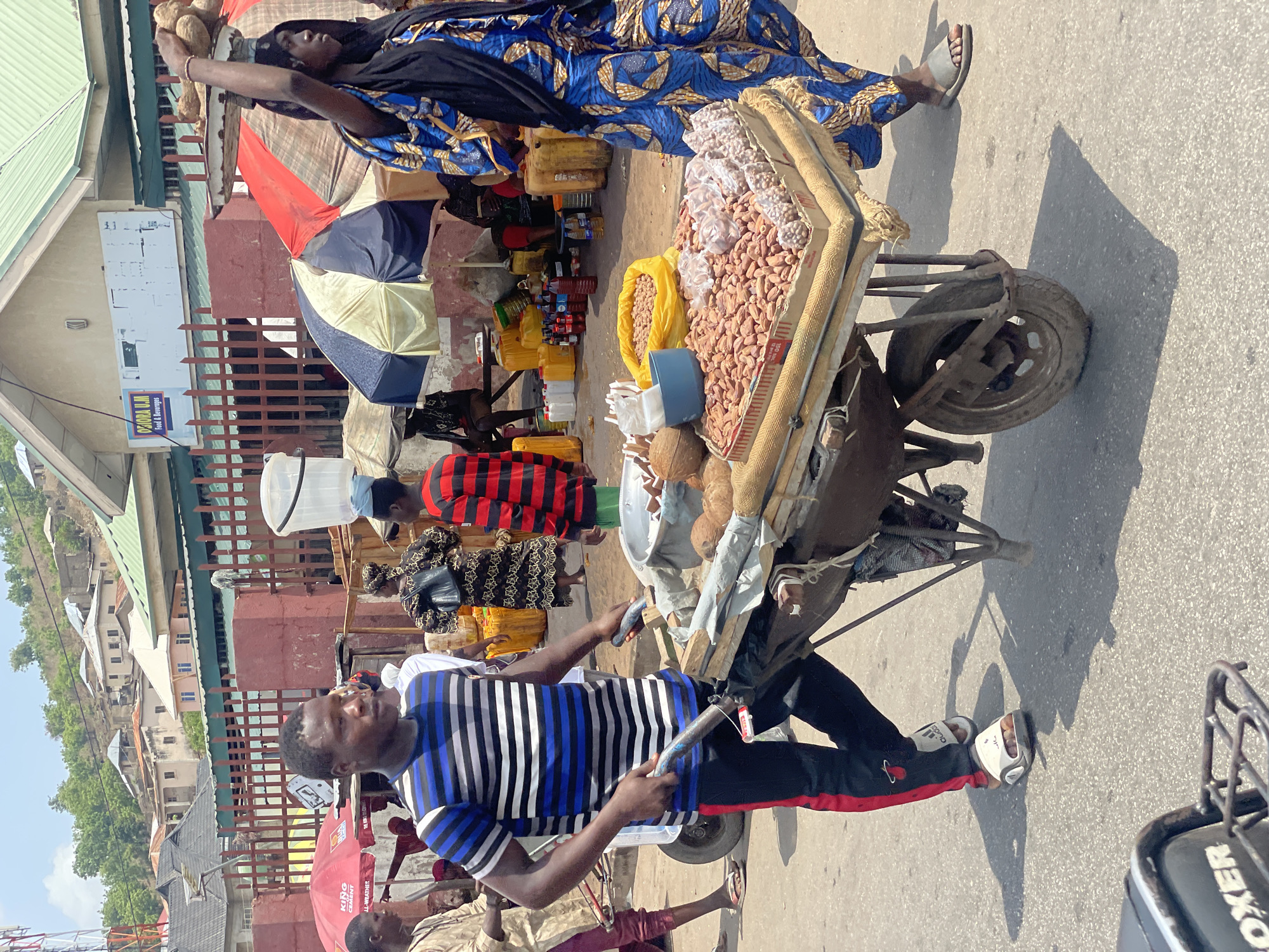 The image depicts a vibrant street scene in Lokoja, Nigeria. A man in a blue and white striped shirt and black track pants pulls a heavily laden cart. The cart is filled with a variety of goods, including peanuts, coconuts, and plastic containers holding various liquids. Above and behind the cart, a woman in a blue patterned dress carries a load of coconuts on her head. Other individuals are visible in the background, some under umbrellas, engaged in commercial activities. The environment features buildings of various heights and designs, typical of an urban area in Nigeria.