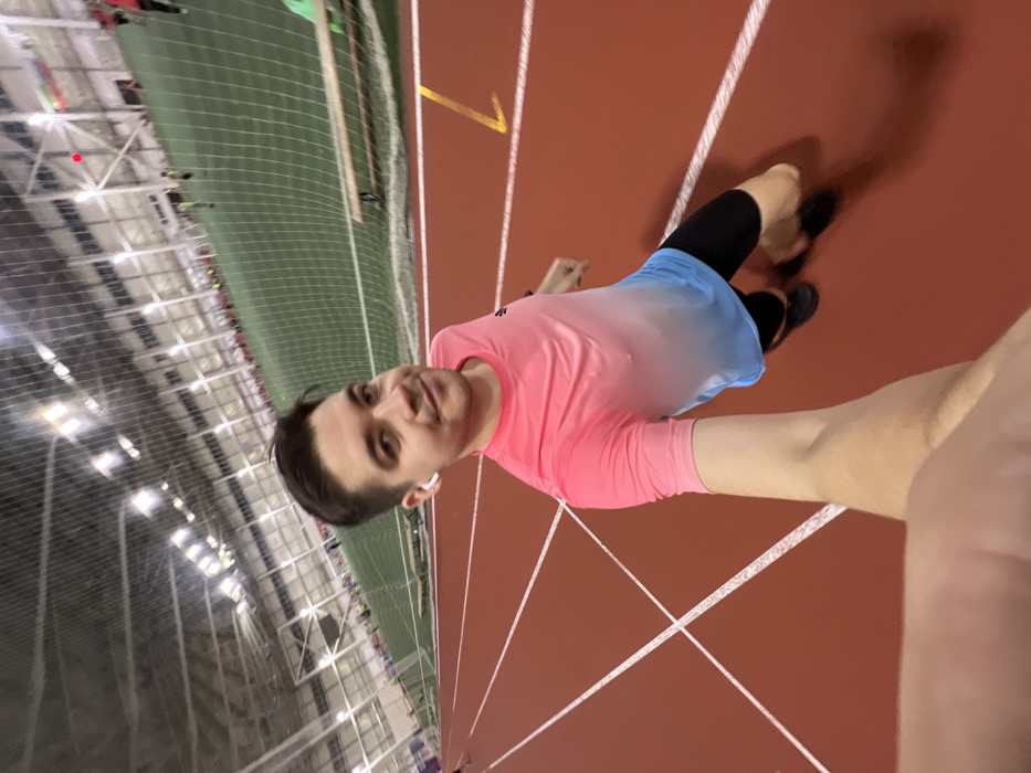 The image displays a young man taking a selfie or being photographed on an indoor running track. The man is positioned in the foreground, looking directly at the camera with a slight smile. He has short dark hair and is wearing a bright pink t-shirt that transitions into a light blue or white gradient towards the bottom, black compression shorts or leggings, and light blue athletic shorts over them. He has white wireless earbuds in both ears. A portion of another person's arm or shoulder is visible in the bottom right corner, suggesting a selfie or close-up shot.
The setting is an indoor athletic facility. The man is standing on a reddish-brown running track with distinct white lane markings. Further down the track, a yellow arrow or symbol is visible. To the left and in the background, a large green artificial turf field, possibly for soccer or football, is visible, enclosed by a protective netting. The ceiling of the facility is high, with numerous bright overhead lights illuminating the space, and the structural beams are also visible. In the distance on the green field, several small, blurry figures can be seen, indicating other people are present at the facility. The man casts a shadow on the track to his right. The overall atmosphere suggests an active sports environment. The image is rotated 90 degrees clockwise.