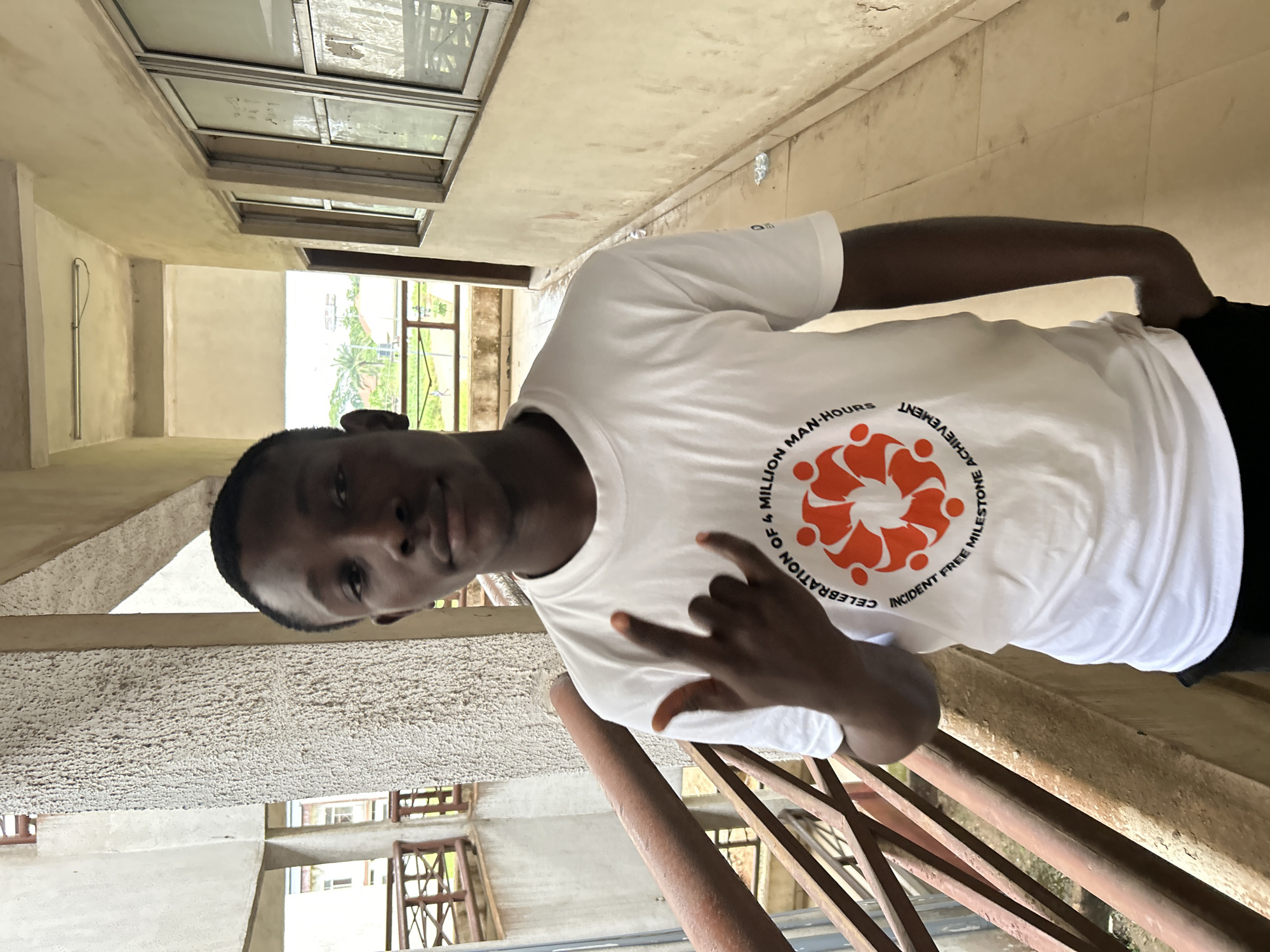 A young Black man, likely in his late teens or early twenties, stands on a metallic staircase, looking towards the camera with a slight smile. He is wearing a white t-shirt with an orange circular logo and text printed on it. The text, partially visible, reads "CELEBRATION OF 4 MILLION MAN-HOURS INCIDENT FREE MILESTONE". The man is making a "rock on" hand gesture with his right hand.
The setting appears to be an outdoor, possibly unfinished, building or hallway. Above and behind the man, there are windows set into a concrete wall. The overall lighting suggests it is daytime, likely bright. The man is positioned on a metal staircase with red-brown railings, and the background shows a textured concrete wall and more windows. The environment seems to be in Ihiagwa, Nigeria. The man's expression is calm and confident.
