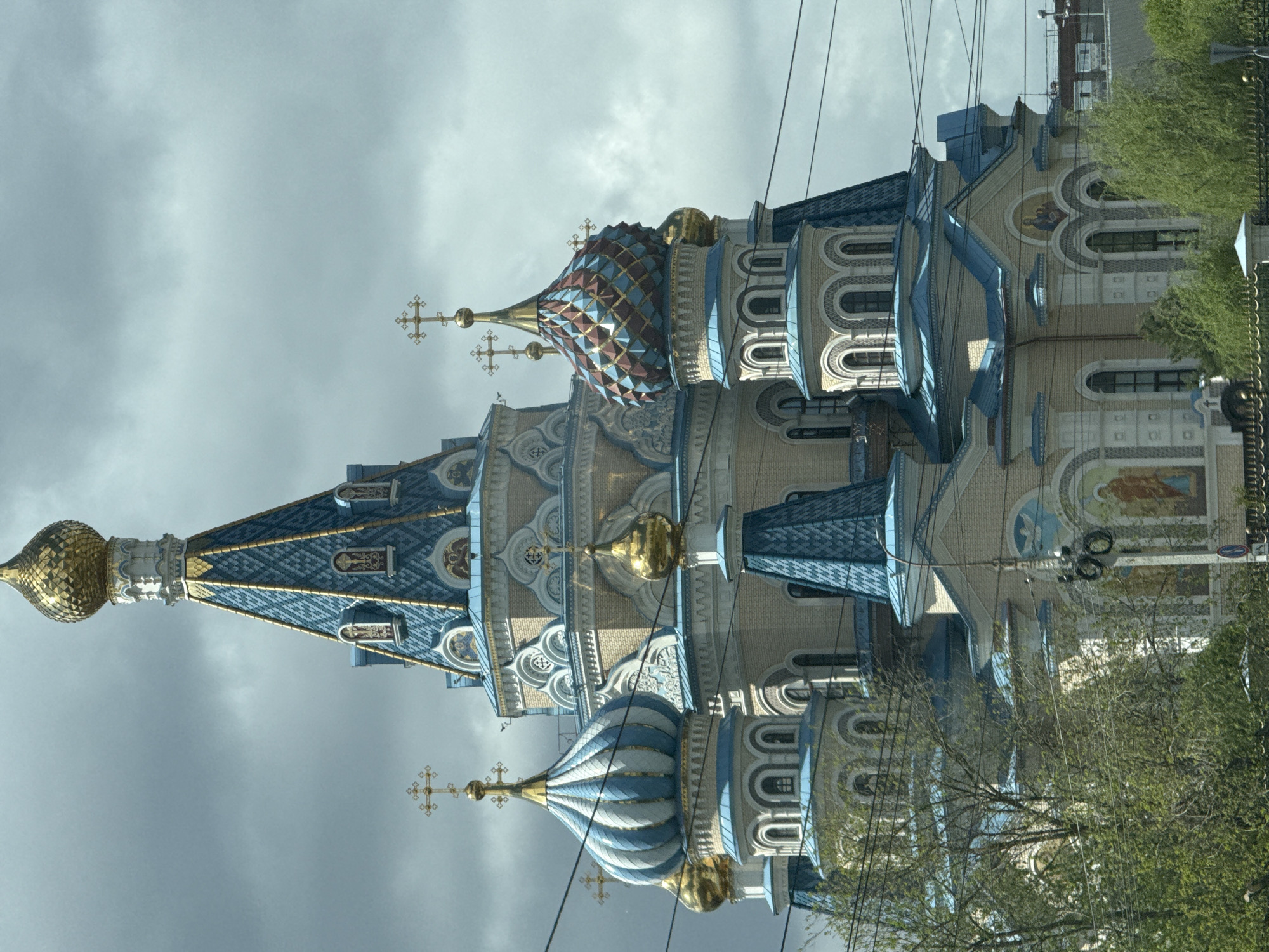The image captures the architectural details of St. Michael's Cathedral in Izhevsk, Russia. The cathedral exhibits multiple domes, each topped with golden crosses. The domes feature intricate patterns and are painted in shades of blue, white, and gold. The structure of the cathedral is a combination of brickwork and ornate stonework with arched windows and decorative elements. Electrical wires cross the sky in the background. In the foreground, tree branches with green leaves are partially visible. The sky is overcast with a muted gray color. A portion of a nearby building can be seen, suggesting the cathedral is located within a city environment.