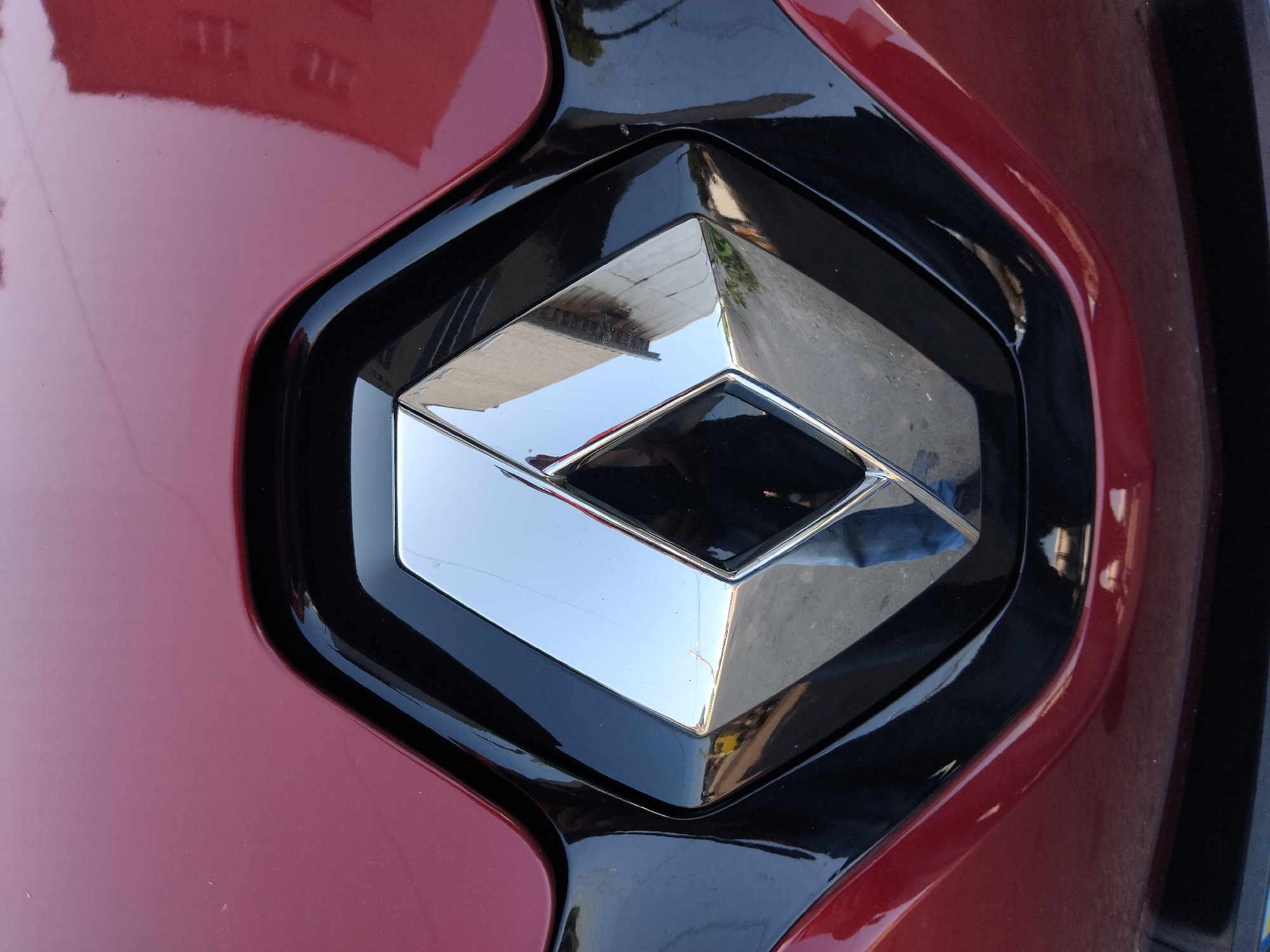 The image shows a close-up of the Renault logo on the front of a dark red car. The logo is a chrome diamond shape with black trim surrounding it, set against the car's bodywork. Reflections are visible in the chrome, showing parts of buildings and what appears to be a person wearing blue jeans. The car is positioned outdoors, with a blurred background that suggests a street or parking area. No specific location in Zhytomyr, Ukraine, can be determined from the image.