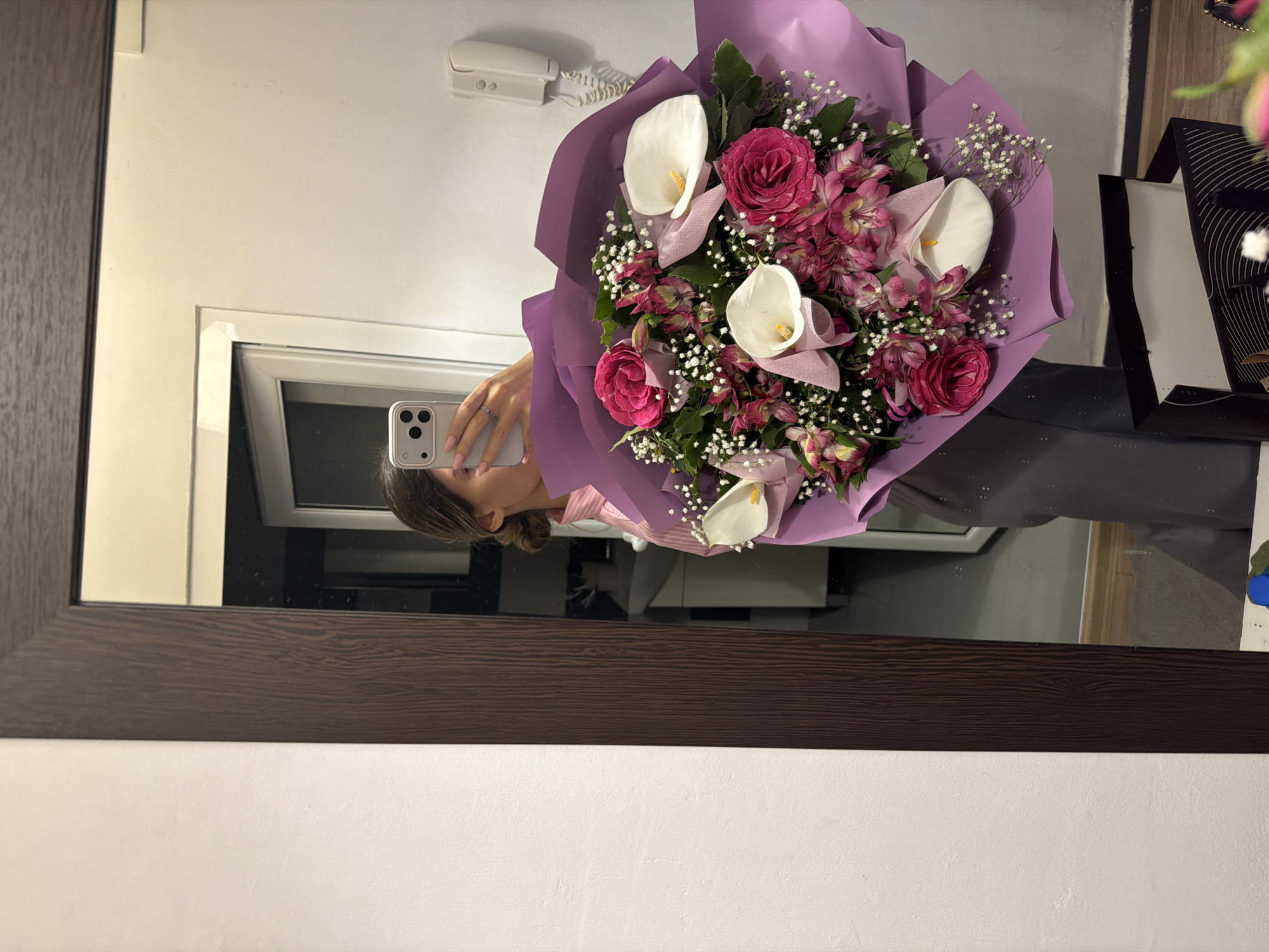 A woman is reflected in a mirror while holding a bouquet of flowers and taking a selfie with her smartphone. The bouquet contains pink roses, white calla lilies, and baby's breath, wrapped in purple paper. A landline telephone is mounted on the wall in the background. The surrounding setting appears to be indoors, possibly in a residential space. There's no discernible information in the image to indicate that it was taken specifically in the city of Sofia, Bulgaria.