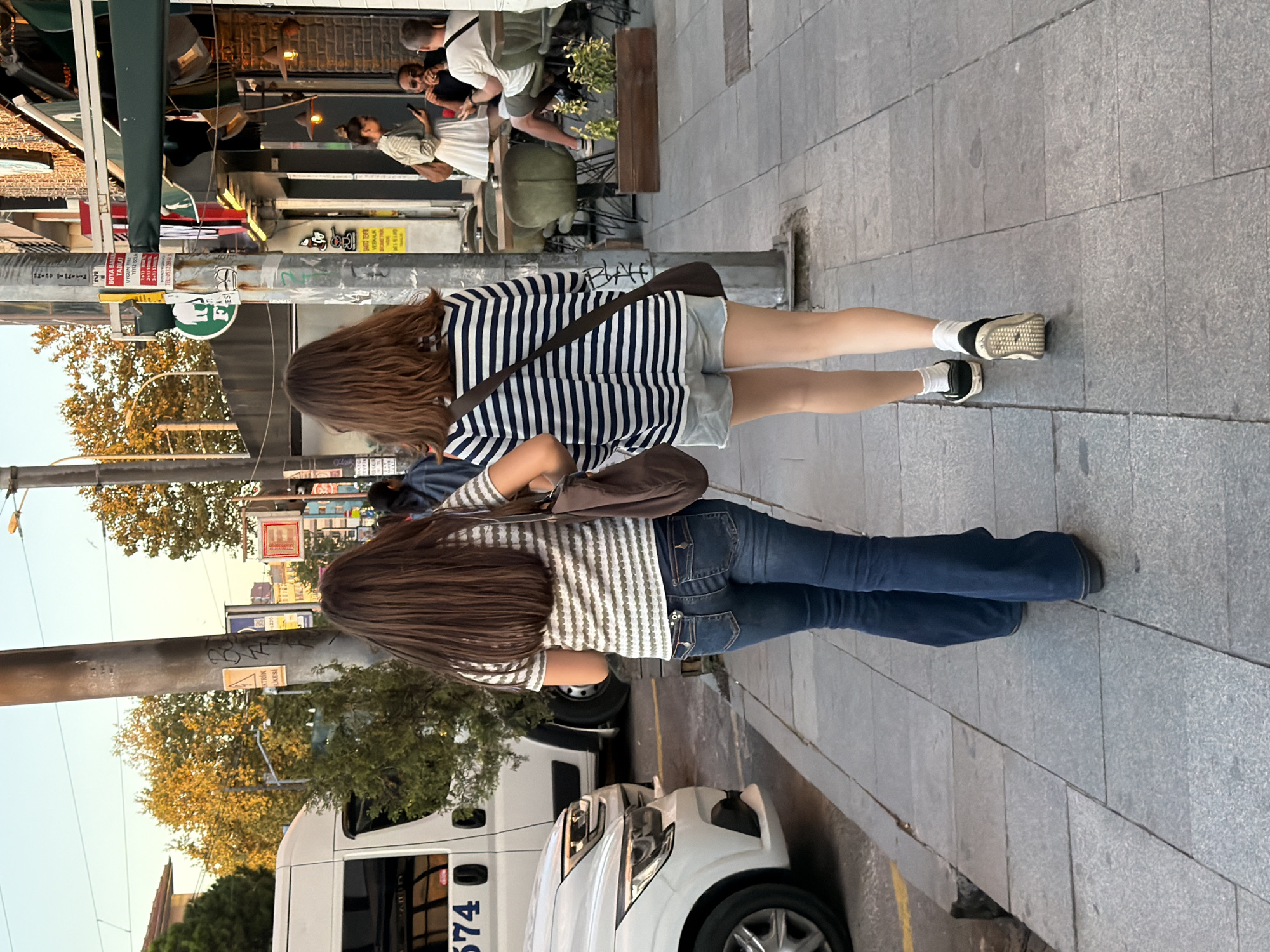 Two young women walk away from the camera on a paved sidewalk in Kadıköy, Turkey. The woman on the left wears a striped t-shirt and flared blue jeans. She carries a brown bag over her right shoulder. The woman on the right wears a horizontally striped long-sleeved shirt and light-colored denim shorts. She also carries a brown bag over her right shoulder and wears black and white sneakers with white socks. Behind them, a white van is parked to their left, and on the right, people sit at tables outside a building with awnings. The scene appears to be a street in a commercial area.