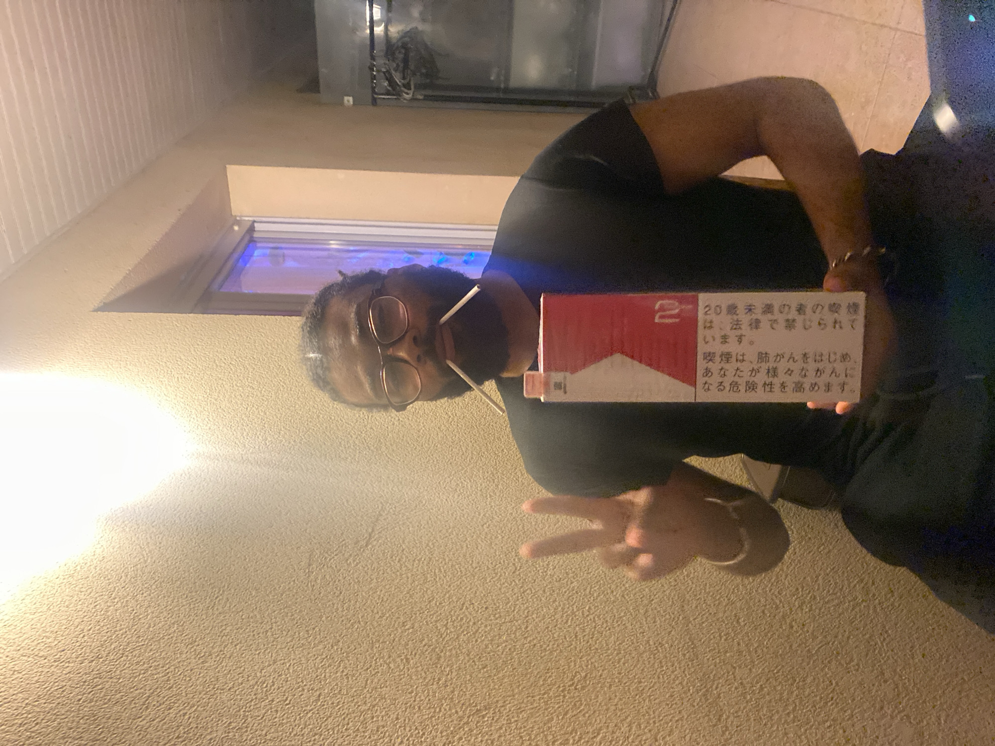 A man, with a dark complexion and wearing glasses, holds a pack of Marlboro cigarettes displaying Japanese text. He is also wearing a dark-colored shirt and is making a peace sign with his hand. The background features a wall with a textured surface, a window with a blue light visible inside, and some kind of metallic equipment. The location appears to be an indoor area, potentially within Montreuil, France.
