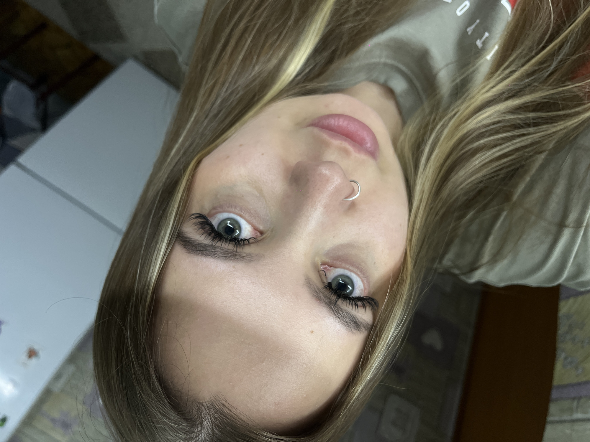 The image shows a Caucasian woman with long, blonde hair and green eyes. She has a silver nose ring in her left nostril. Her eyebrows are dark and full, and she has eyelash extensions. She is wearing a light-colored t-shirt. The background is blurry and difficult to discern, but it appears to include a white surface and some furniture. It is not possible to determine the location in Port-Katon, Russia from the image.