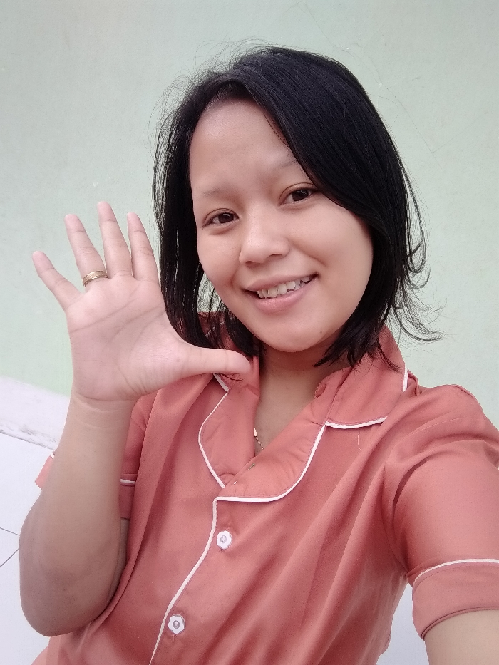 A smiling woman poses indoors against a light green wall. She raises her left hand, displaying a gold ring on her ring finger. The woman has short, dark hair and fair skin. She wears a coral-colored, short-sleeved shirt with white piping along the collar and button placket. White buttons are visible. The light suggests the photo was taken during the day.