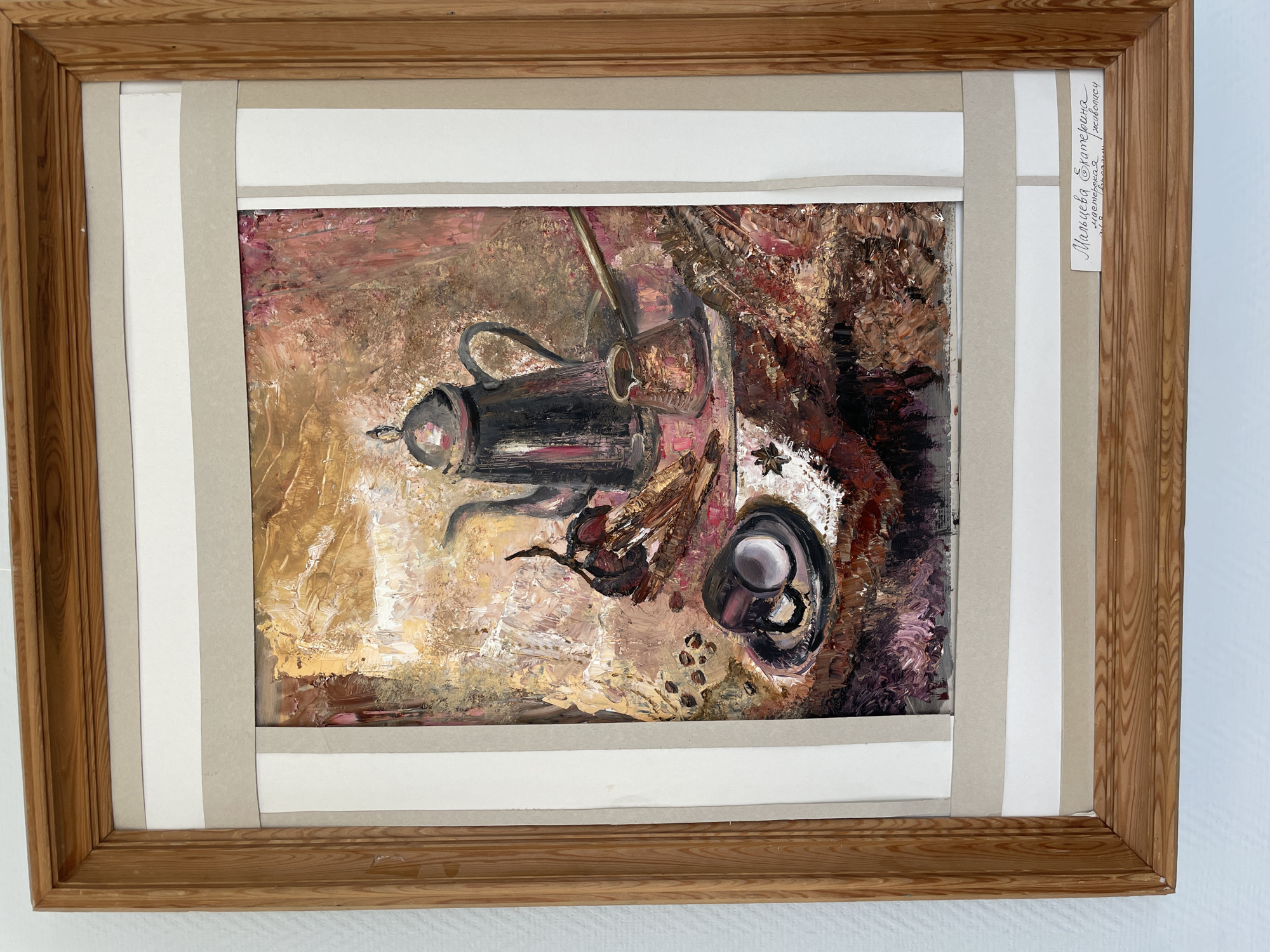 This is a photograph of an oil painting framed in a light-colored wooden frame. The painting depicts a still life with a dark, metallic coffee pot with a curved handle, a small cup on a saucer, and scattered coffee beans. A spice, possibly star anise, is also visible on the surface. The background is textured with earthy tones of brown, gold, and red, suggesting a rustic or warm setting. The brushstrokes are thick and visible, giving the painting a rich, impasto texture.
Attached to the right side of the frame, a small white label contains handwritten text in Cyrillic script. The text appears to be a signature or title, possibly indicating the artist's name and the artwork's title. The handwriting suggests a personal or artisanal touch.
The overall impression of the painting is one of quiet contemplation, perhaps a moment of enjoying a warm beverage in a cozy setting. The colors and textures evoke a sense of warmth and comfort. Given the location context of Perm, Russia, the scene could represent a common domestic or cafe setting in the region. The lighting suggests it is indoors, possibly during the daytime, but the focus is on the still life itself rather than the external environment.