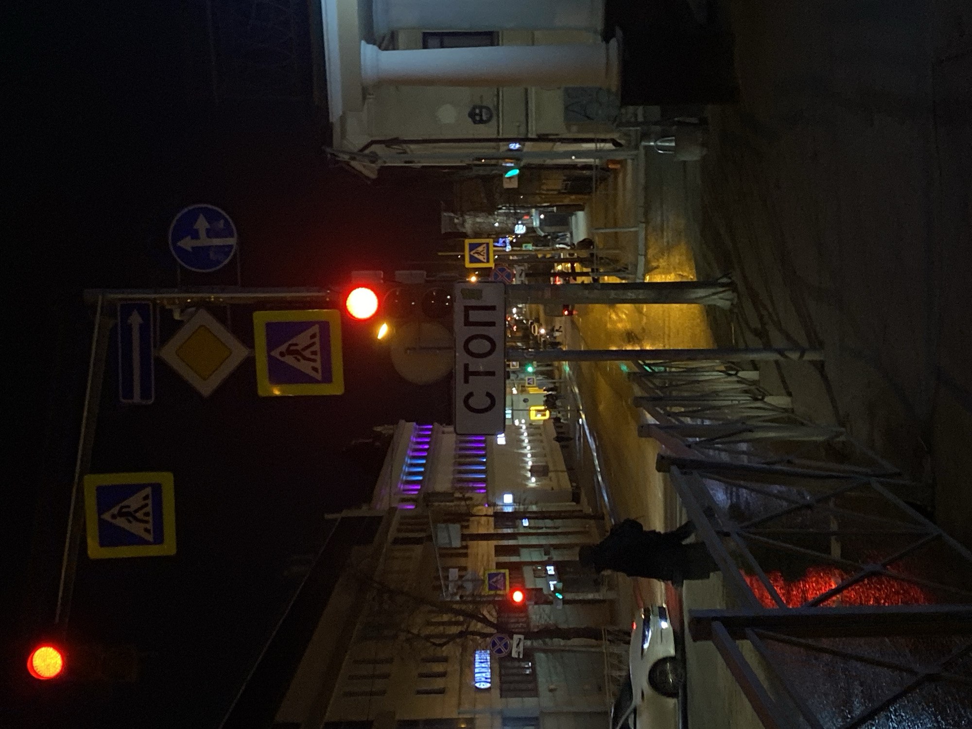 The image shows a night scene in what appears to be the city of Kazan, Russia, evident from the Cyrillic lettering on the "STOP" sign. The view is of a street with multiple pedestrian and directional signs along with traffic lights, currently showing red. There is a building illuminated with purple lighting on one side of the street. A person, possibly a pedestrian, is walking along the sidewalk behind a metal railing. A white vehicle is parked on the side of the road near the railing. The street appears wet, likely due to recent rain or cleaning. The overall lighting is dim, with the streetlights and illuminated signs and buildings providing the main sources of light.